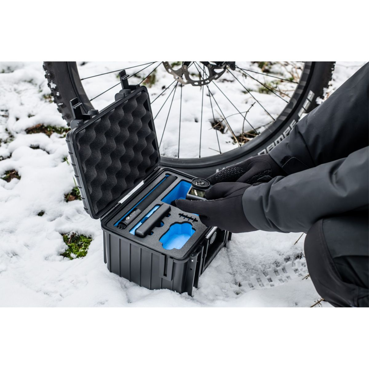 Person in Handschuhen öffnet eine schwarze Kiste mit Ausrüstung neben einem Fahrradreifen im Schnee.
