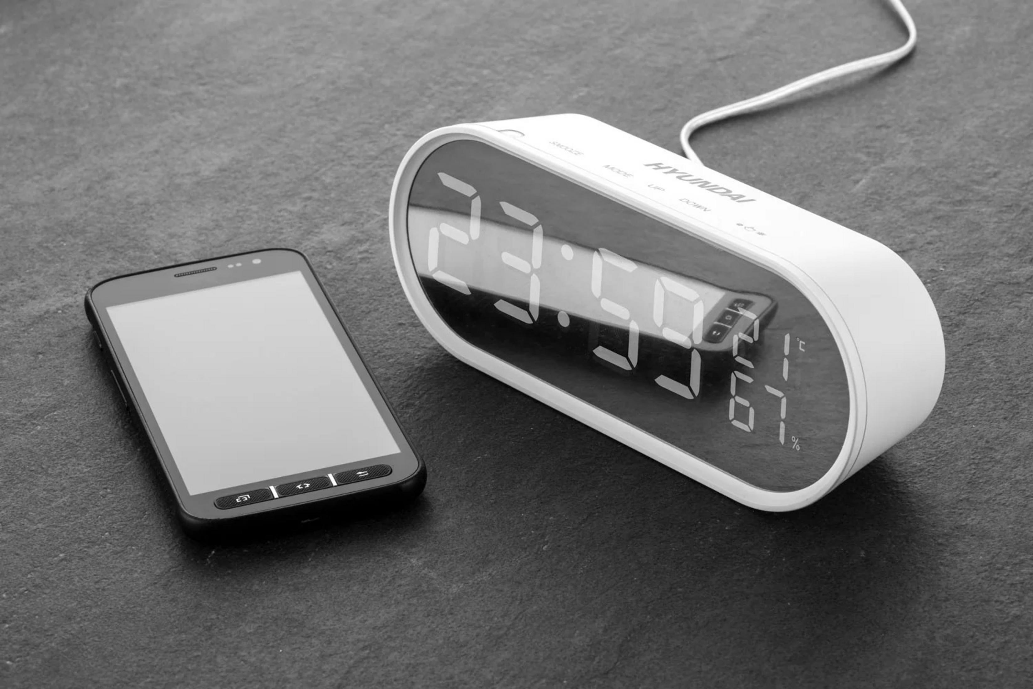 Czarny smartfon leży obok białego cyfrowego budzika na ciemnym kamieniu.