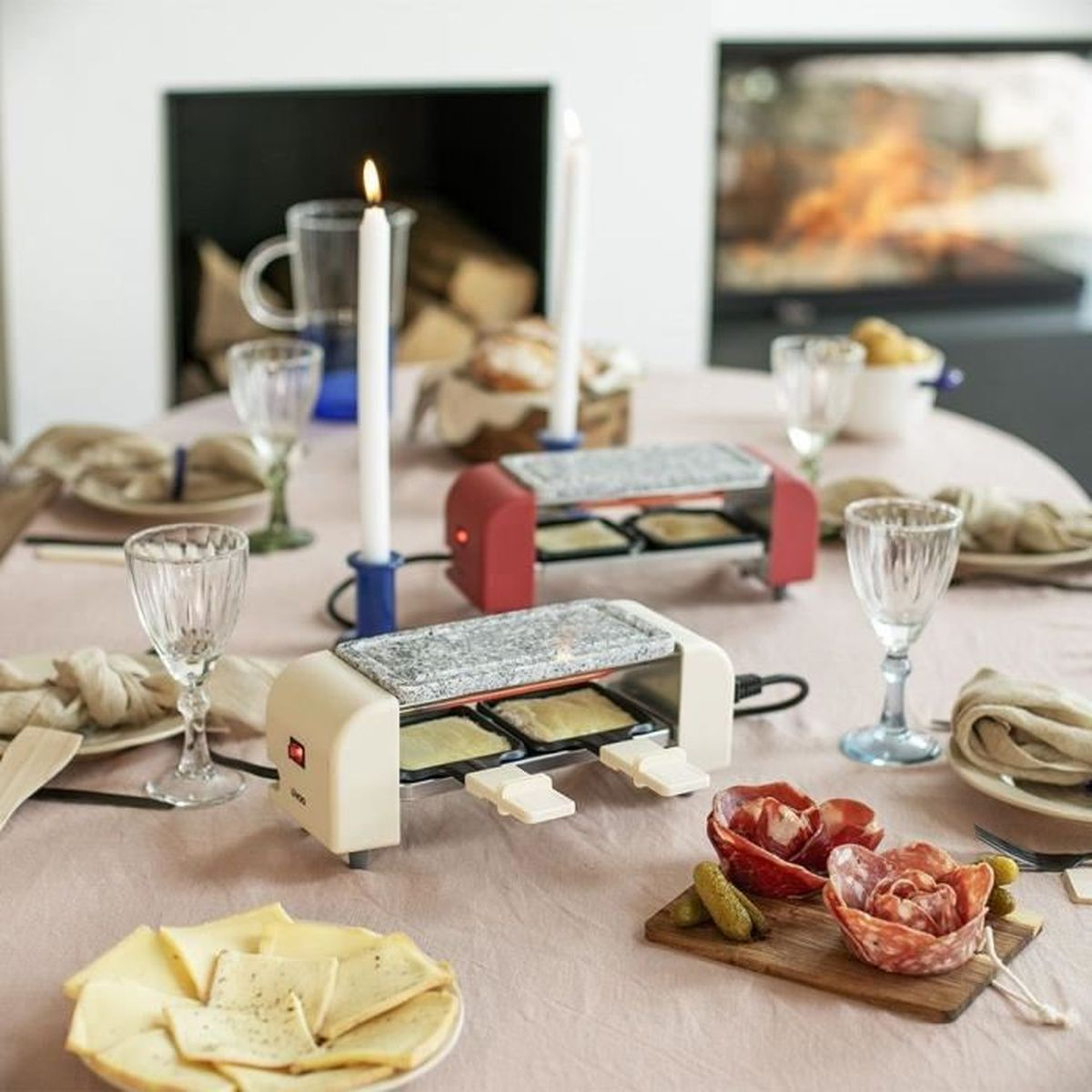Ein Tisch ist für ein Raclette-Dinner gedeckt, mit zwei Raclette-Öfen, Kerzen und Käse.