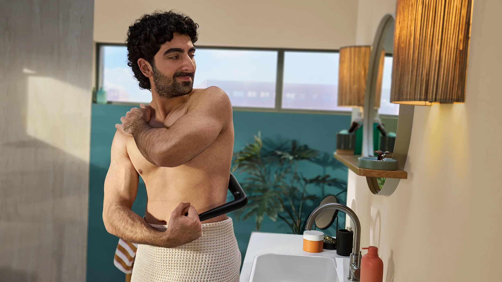 Ein Mann mit dunklen Locken rasiert seinen Rücken im Badezimmer mit einem Handtuch um die Hüften.