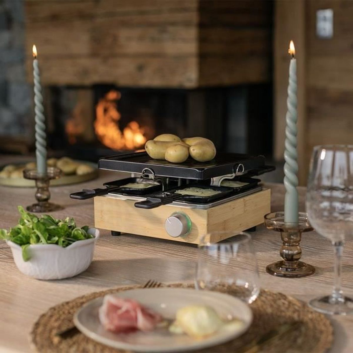 Raclette-Grill mit Kartoffeln und Käse, Kerzen und Kamin im Hintergrund.