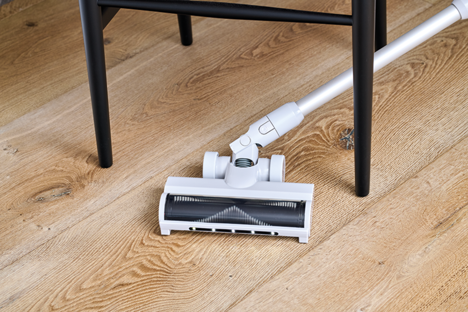 Tête d'aspirateur blanche avec brosse sur parquet, à côté d'un pied de chaise noir.