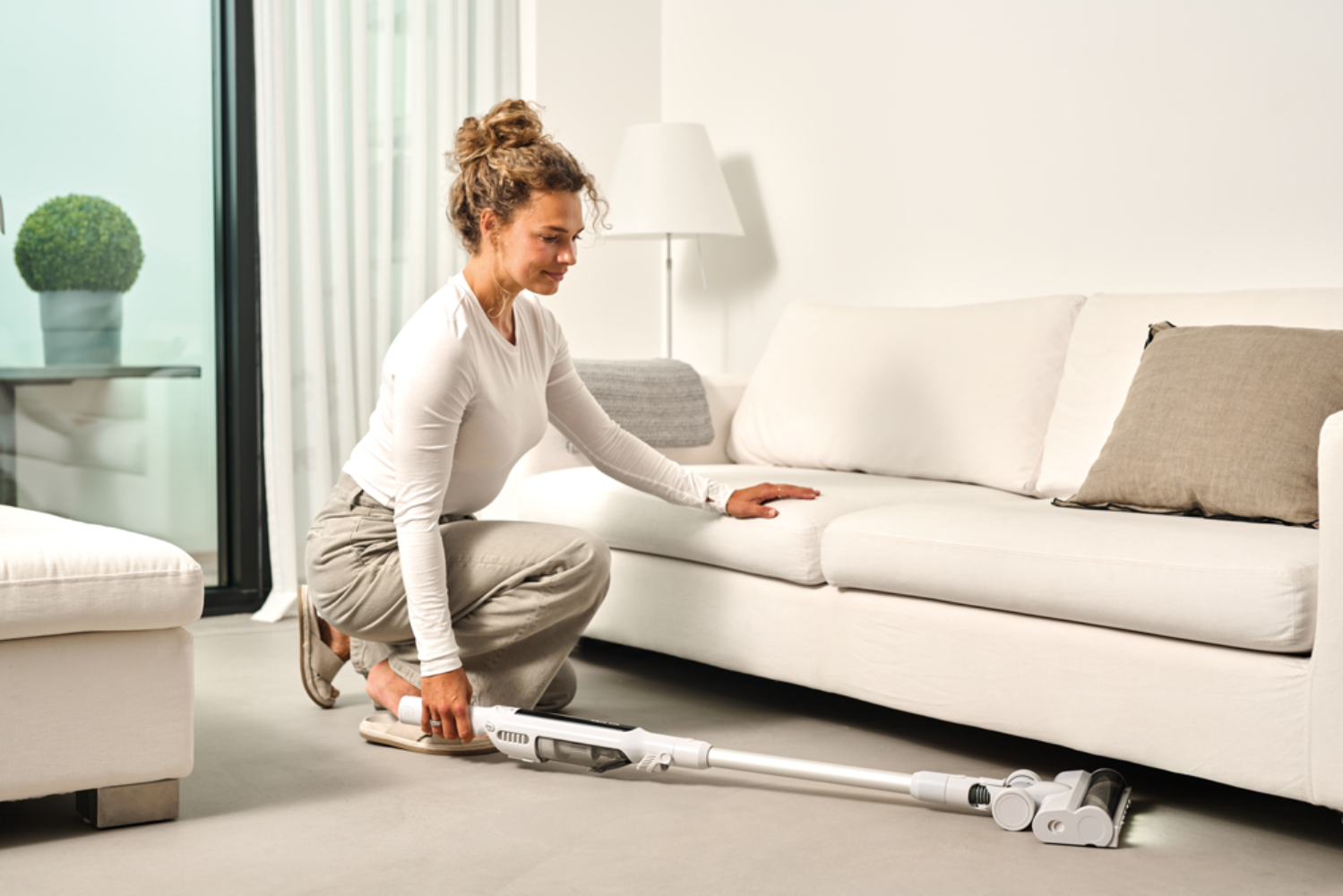 Une femme s'agenouille au sol utilisant un aspirateur blanc sous un canapé blanc.
