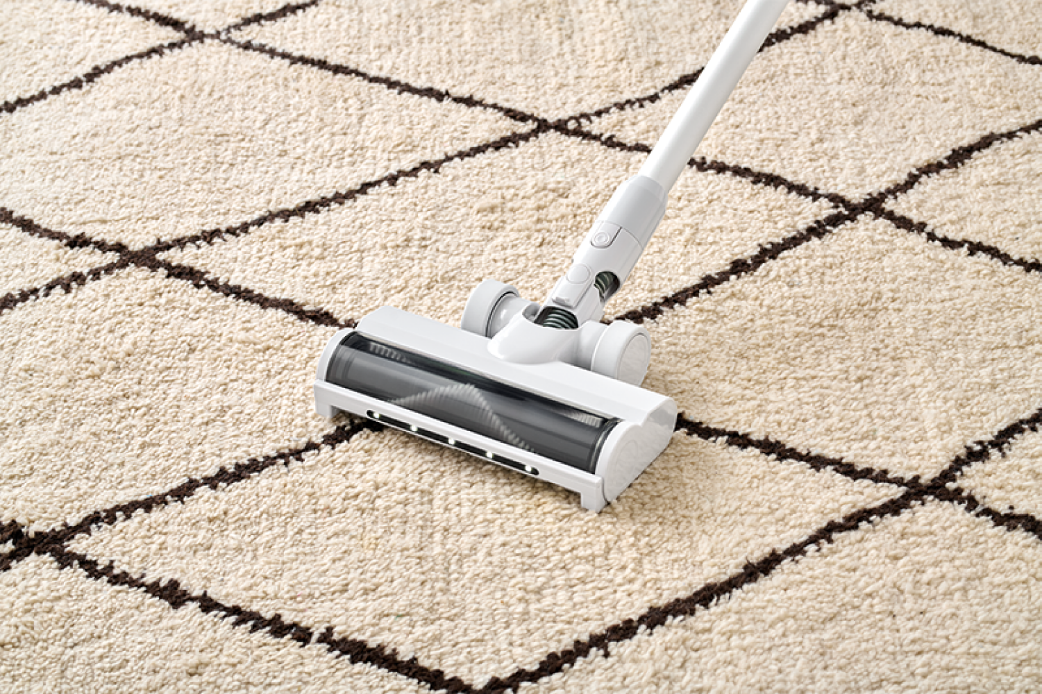 Un aspirateur blanc nettoie un tapis crème à motifs losanges marron.