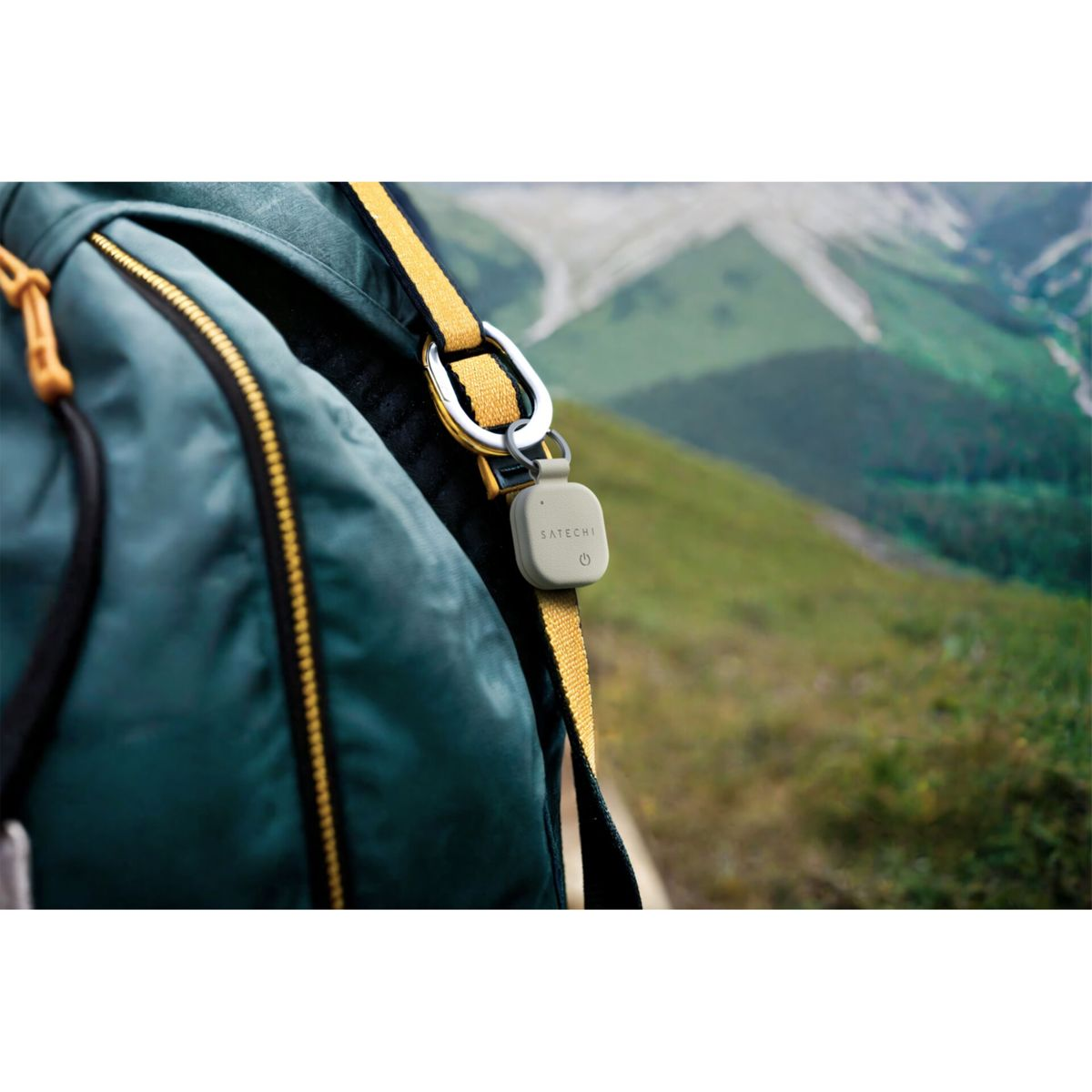 Ein grauer Tracker hängt an einem Rucksack, im Hintergrund grüne Berge.