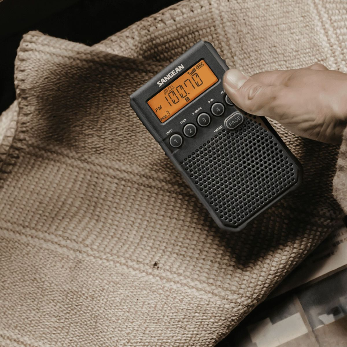 Una mano tiene una radio Sangean nera con display arancione. Appoggiata su tessuto.