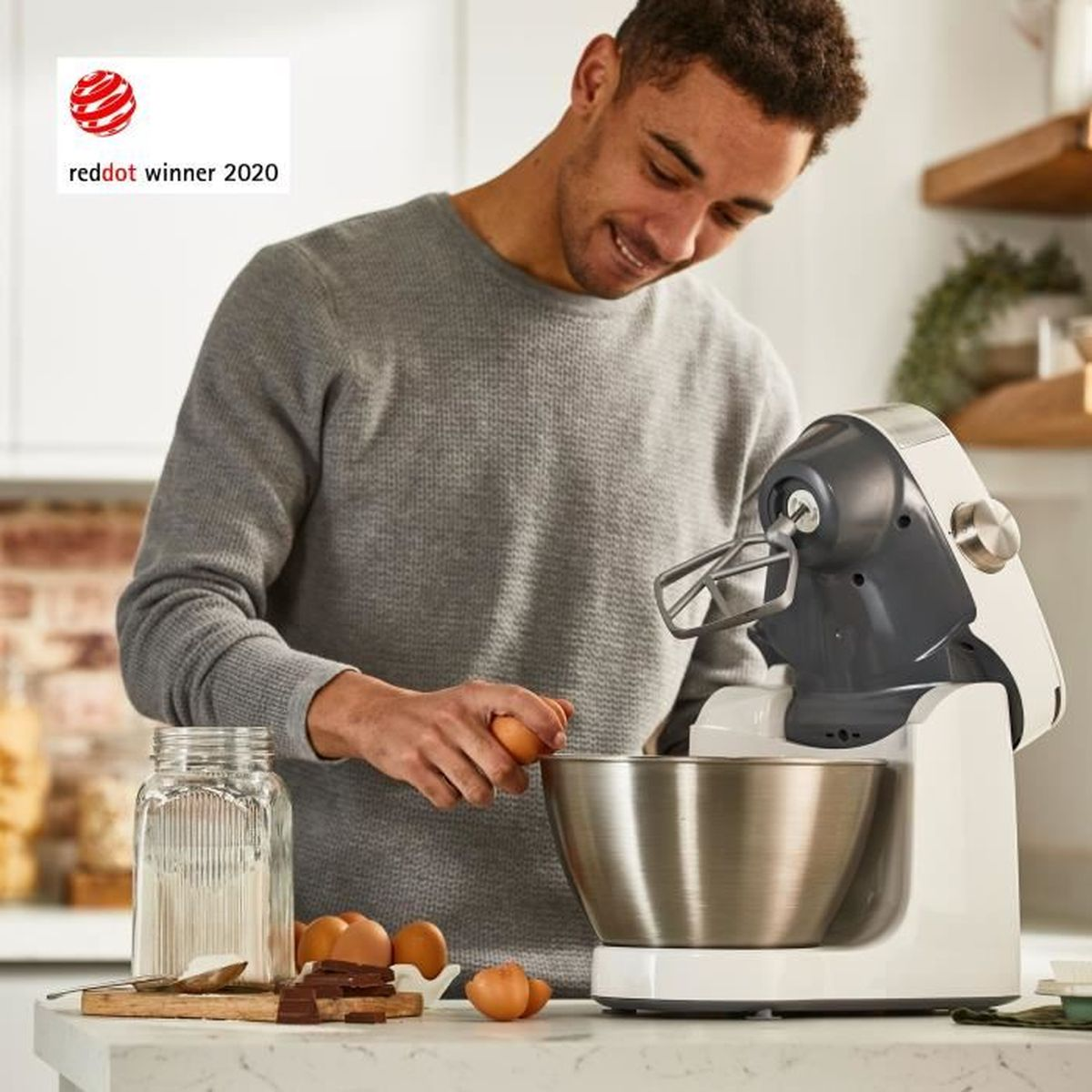 Mann gibt Eier in Küchenmaschine. Glas, Schokolade, Zutaten auf Tisch.