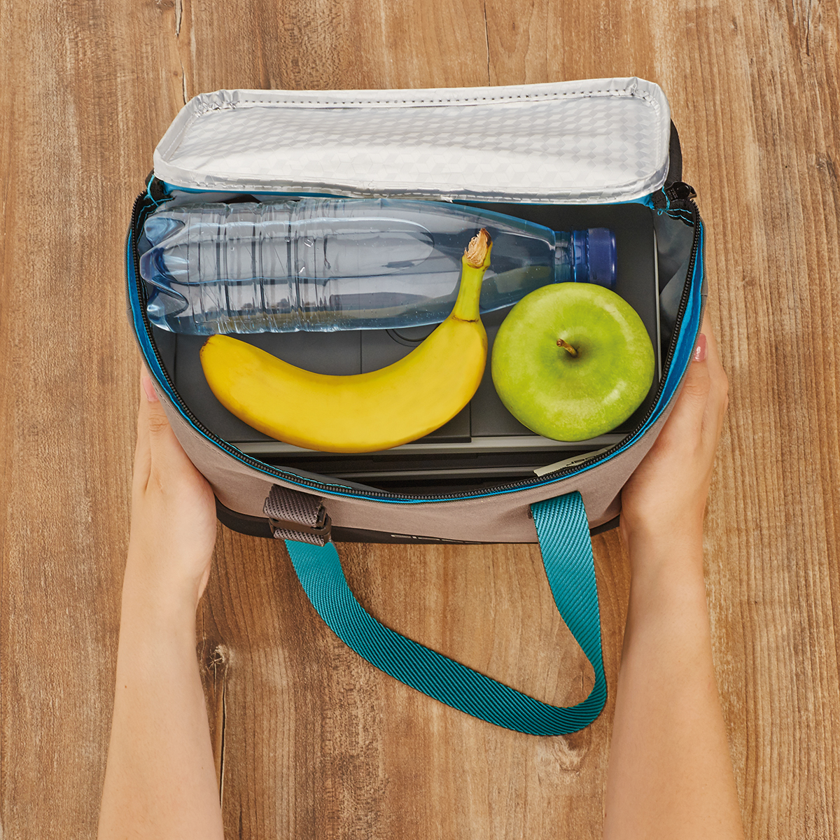 Hände halten eine Lunchtasche offen mit Wasserflasche, Banane und grünem Apfel.
