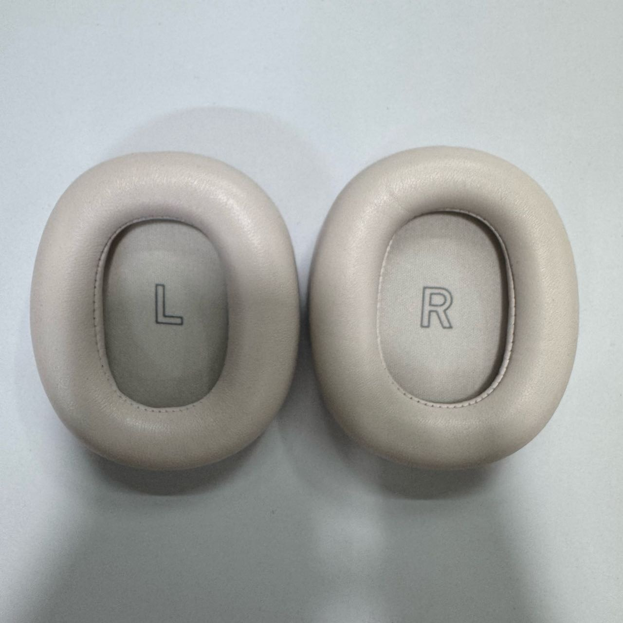 Zwei beige Ohrpolster nebeneinander, eines mit 'L', das andere mit 'R'.