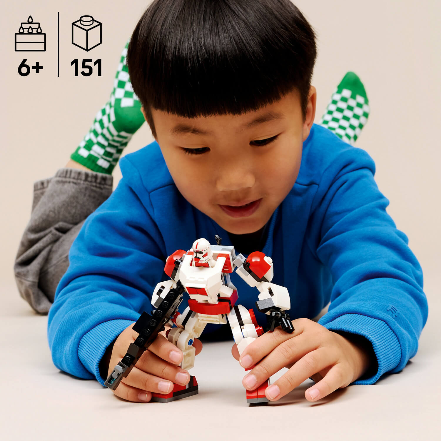 Bir erkek çocuk karnının üzerinde yatıyor, kırmızı ve beyaz Lego robotla oynuyor.