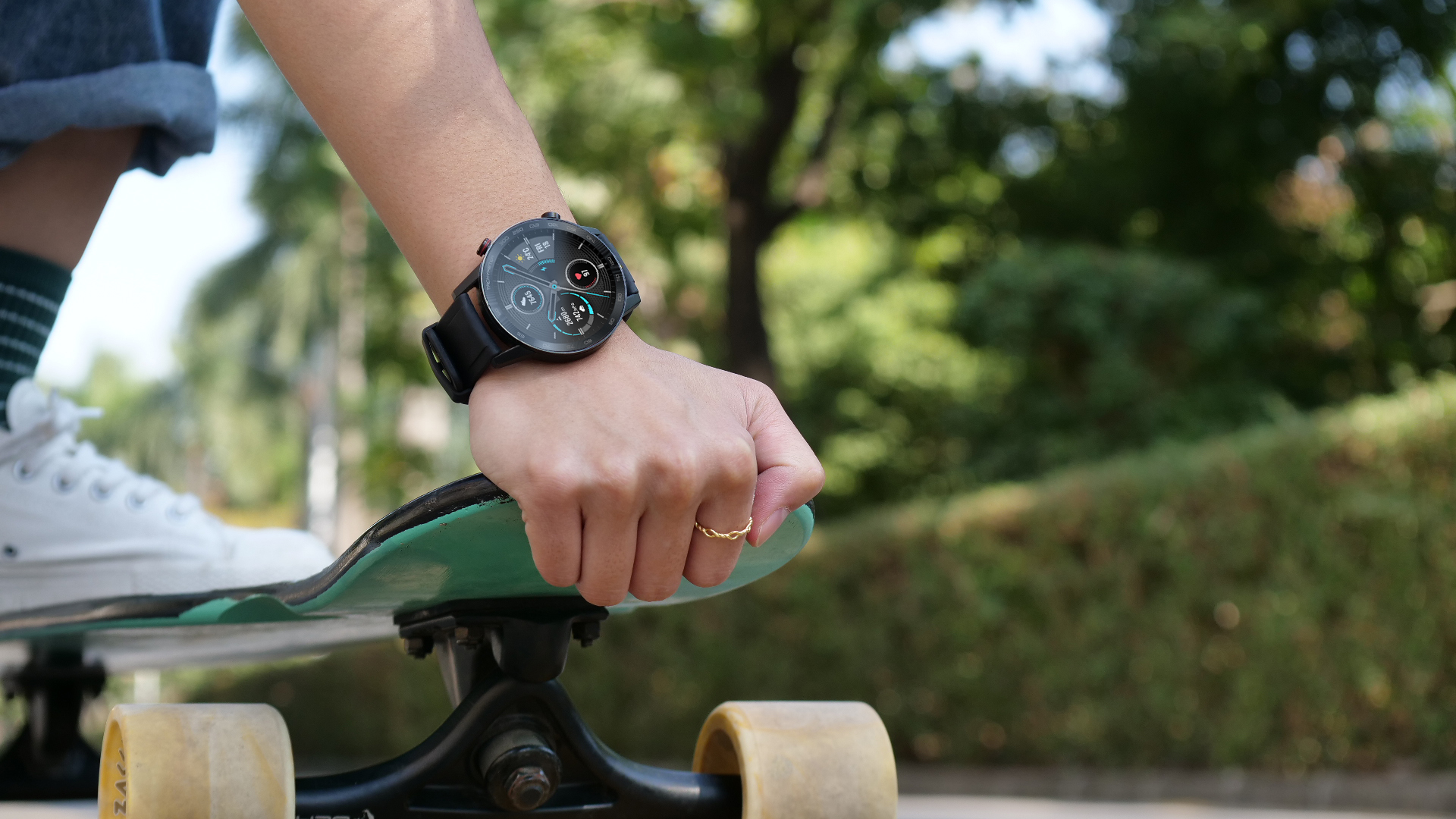 Eine Hand mit Smartwatch und Ring hält ein Skateboard. Weiße Schuhe und grüne Socken sichtbar.