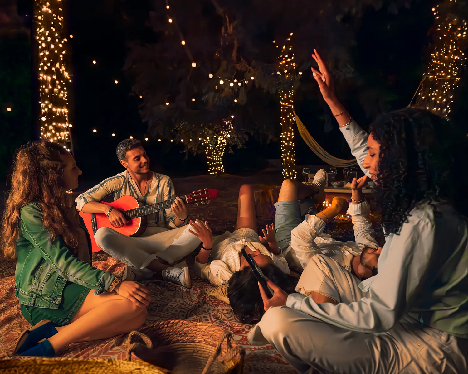 Przyjaciele siedzą na zewnątrz w nocy, oświetleni lampkami, jeden gra na gitarze.