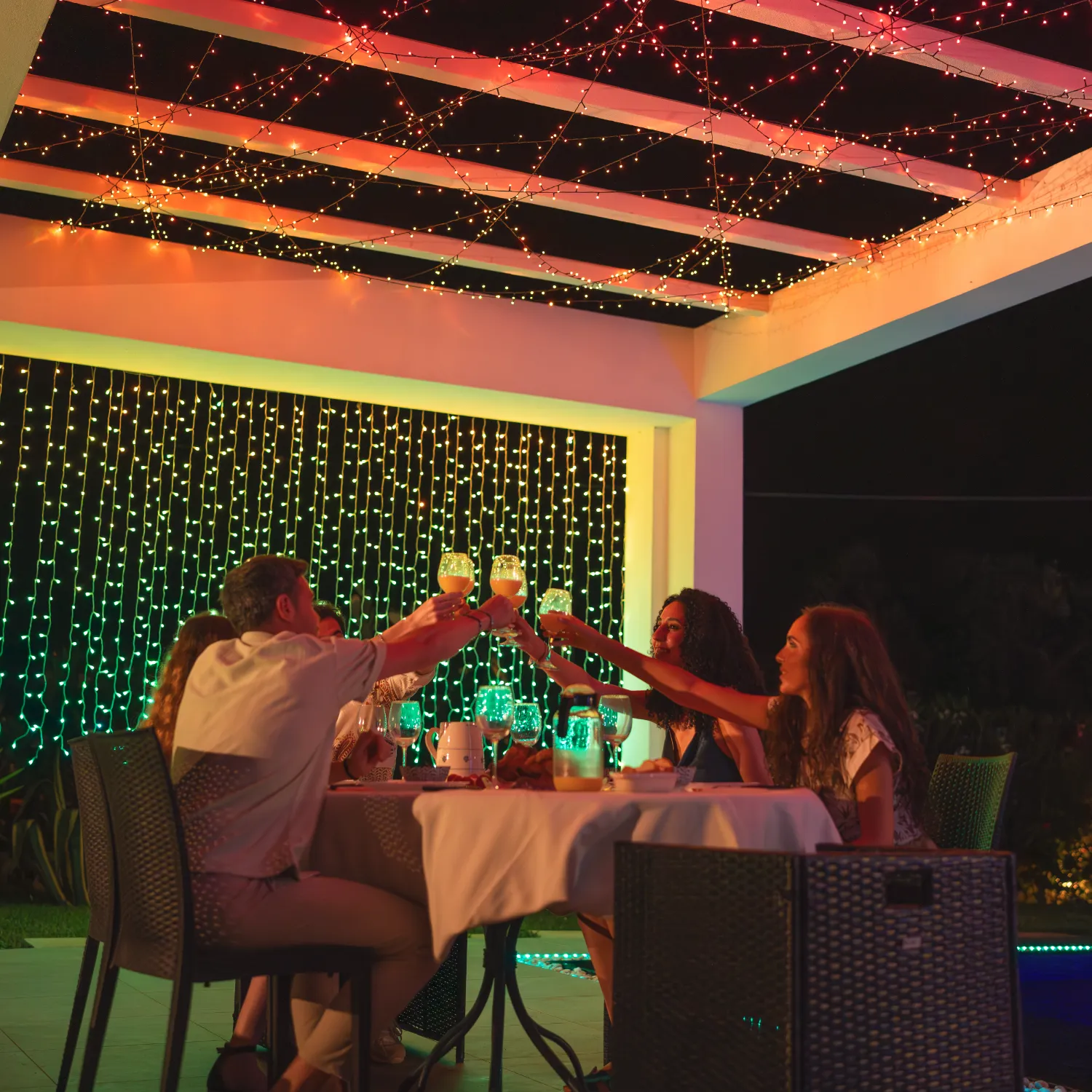 Ludzie wznoszą toasty napojami pod kolorowymi lampkami na patio nocą.