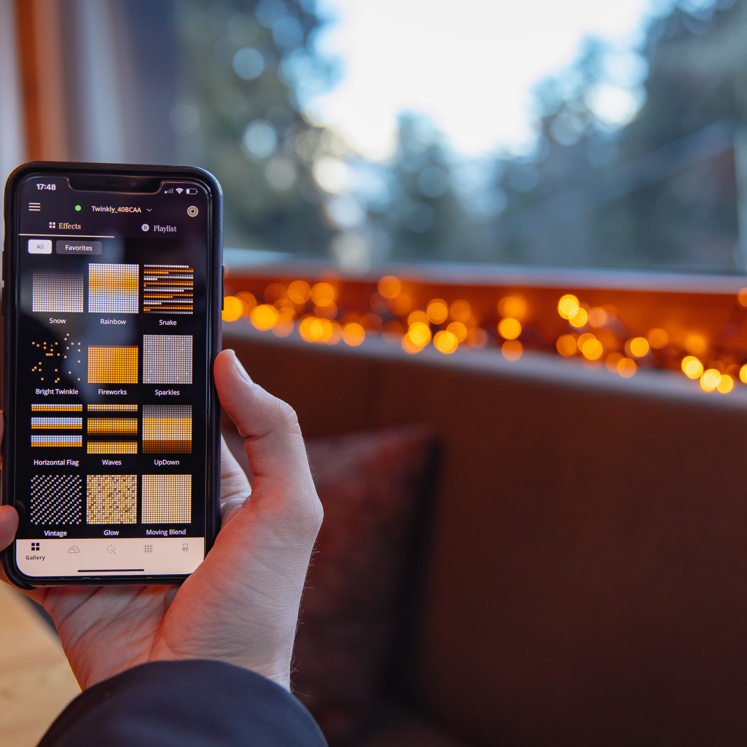 Ręka trzyma smartfon z aplikacją Twinkly, pokazuje efekty świetlne. Pomarańczowe lampki w tle.
