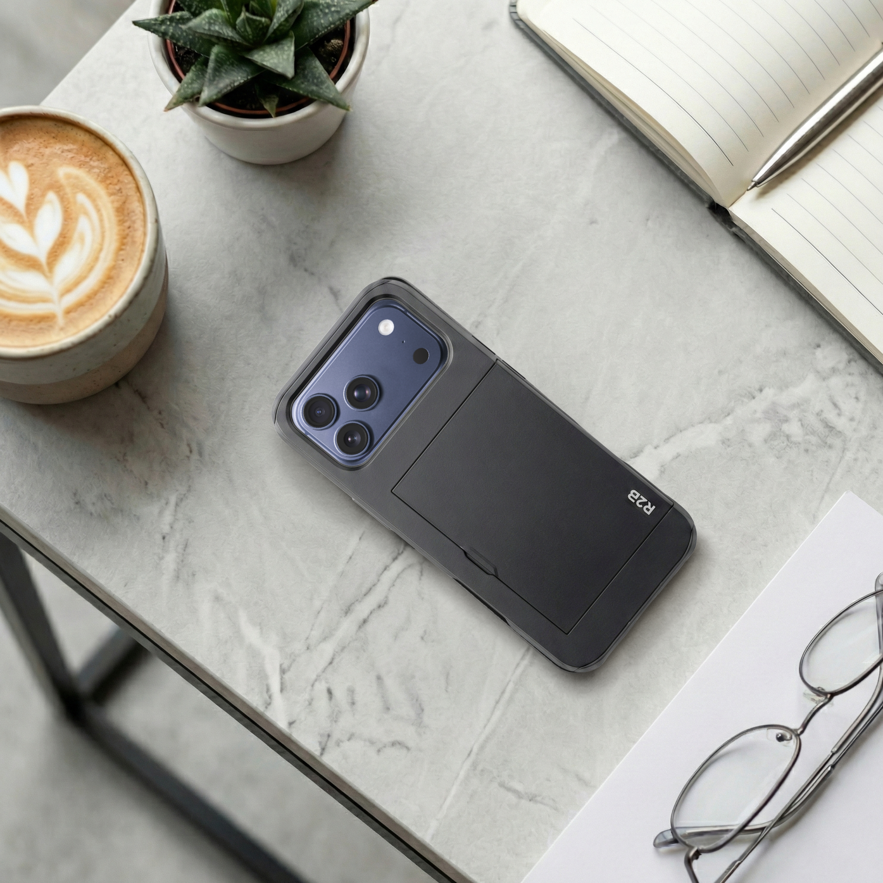Une coque de téléphone noire avec objectifs, café, plante et carnet sur table en marbre.