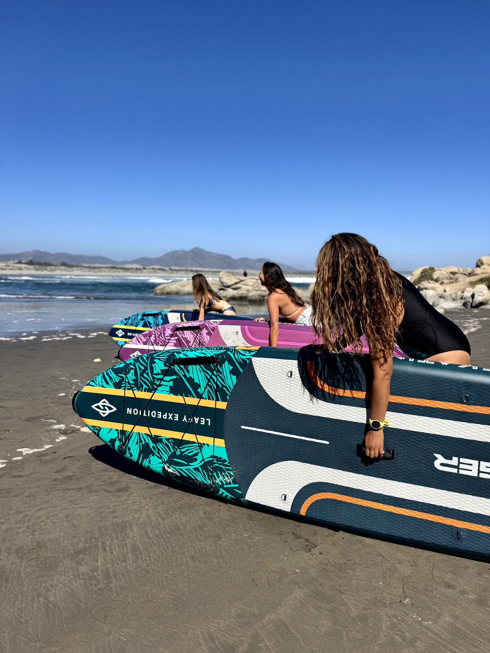 Trzy kobiety leżą na desekach do paddleboardingu na plaży, z morzem w tle.