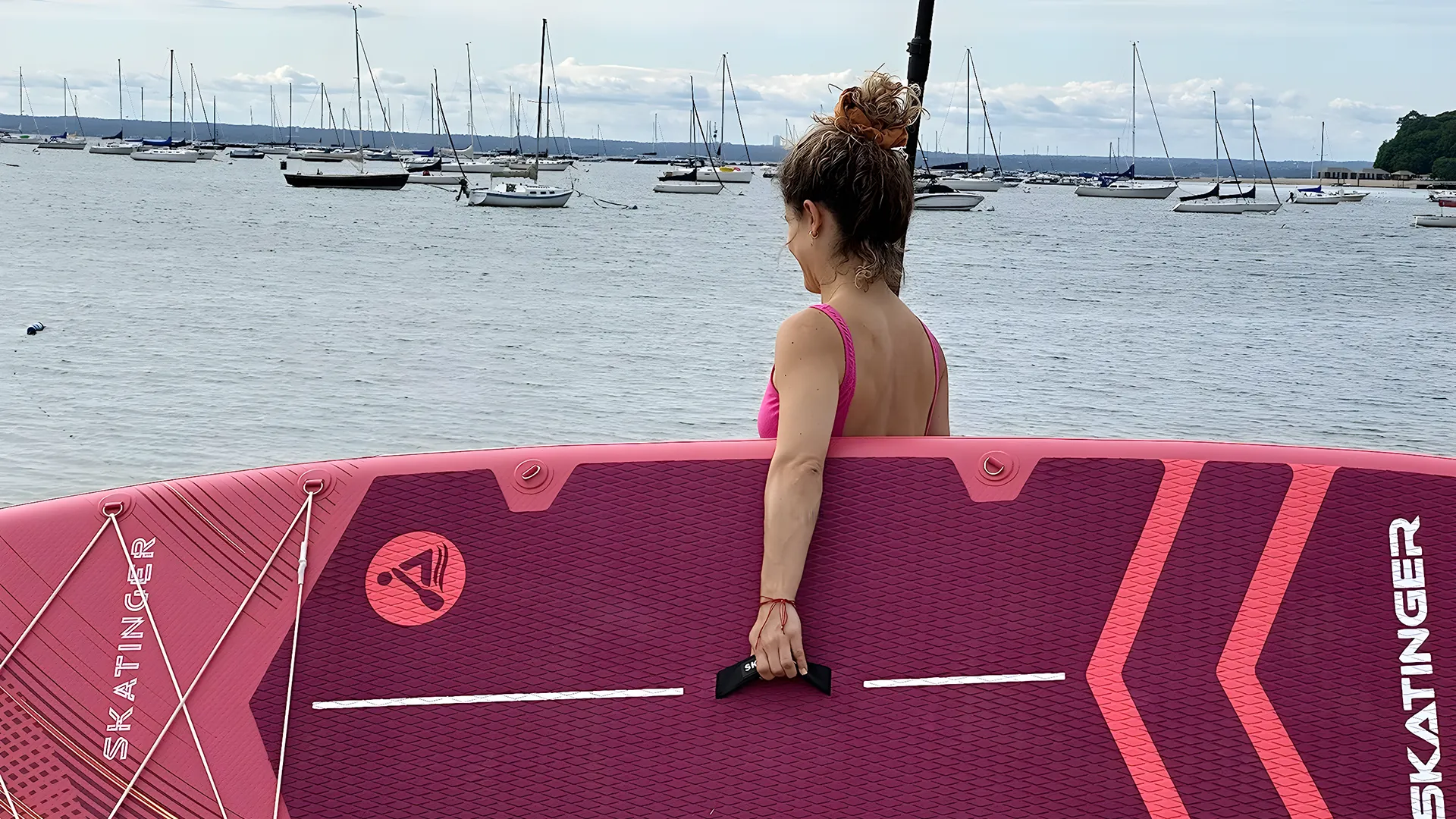 Kobieta w różowym bikini trzyma różową deskę SUP. W tle wiele żaglówek.