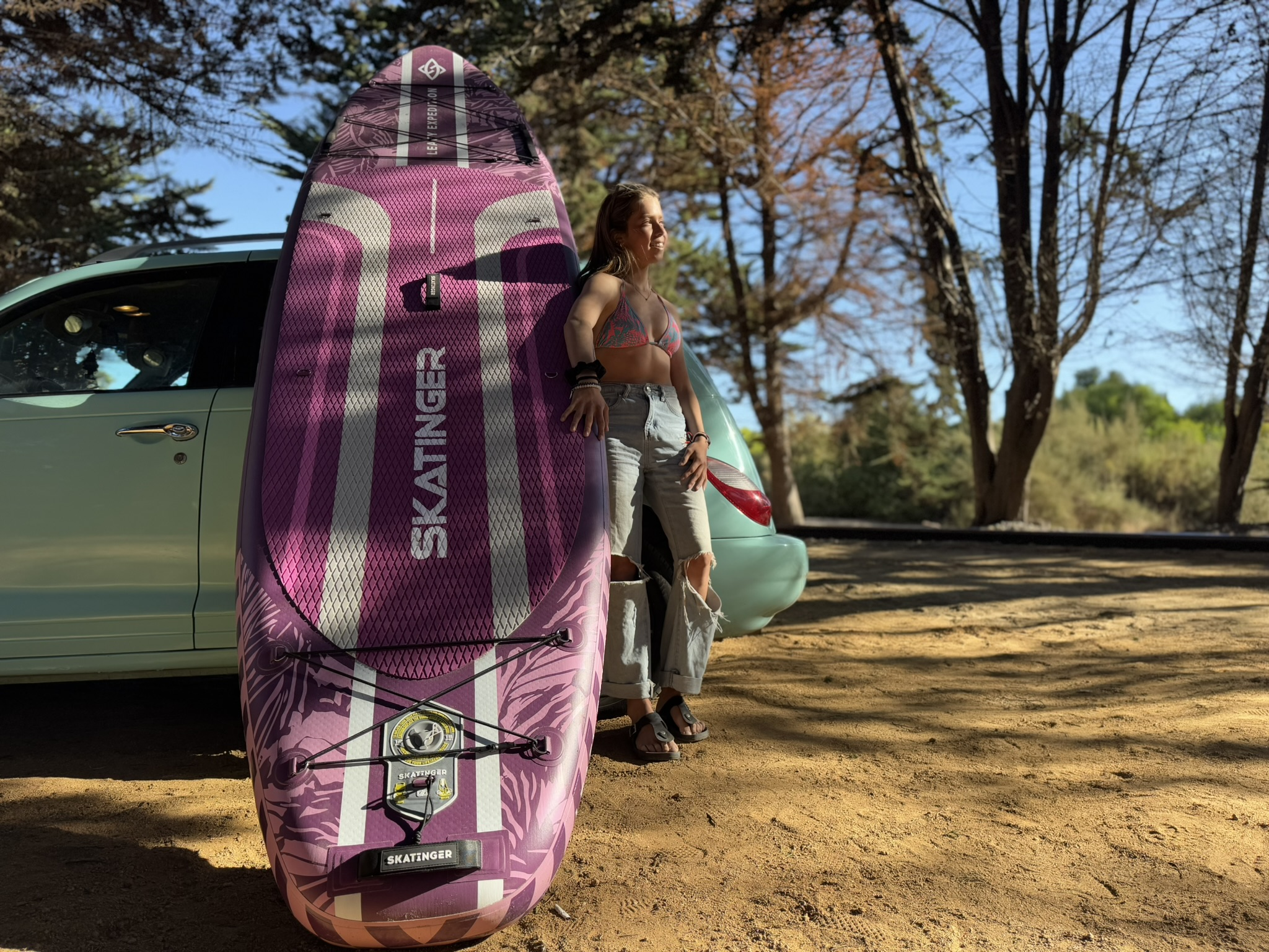 Kobieta opiera się o fioletową deskę surfingową obok samochodu, drzew i błękitnego nieba.