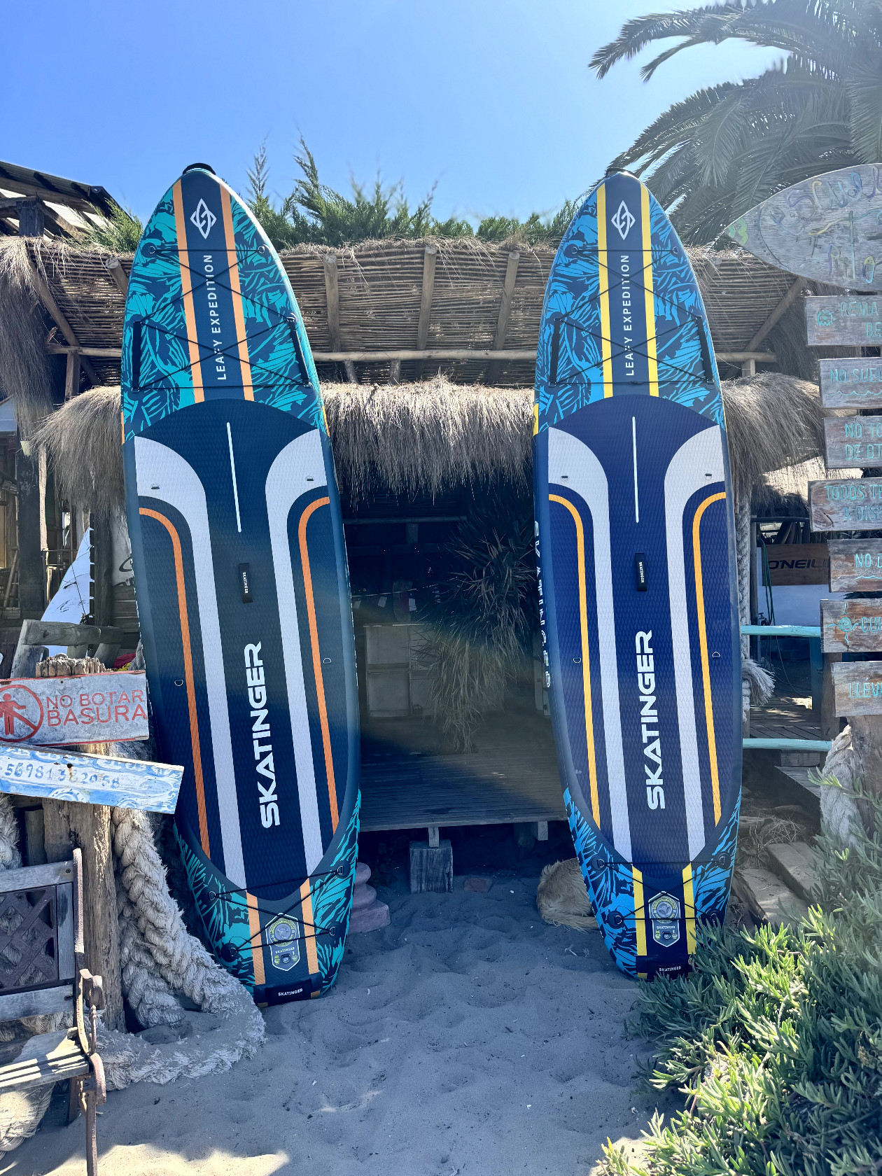 Dwie deski surfingowe stoją na piasku przed chatką z dachu krytego strzechą. Czyste niebo.