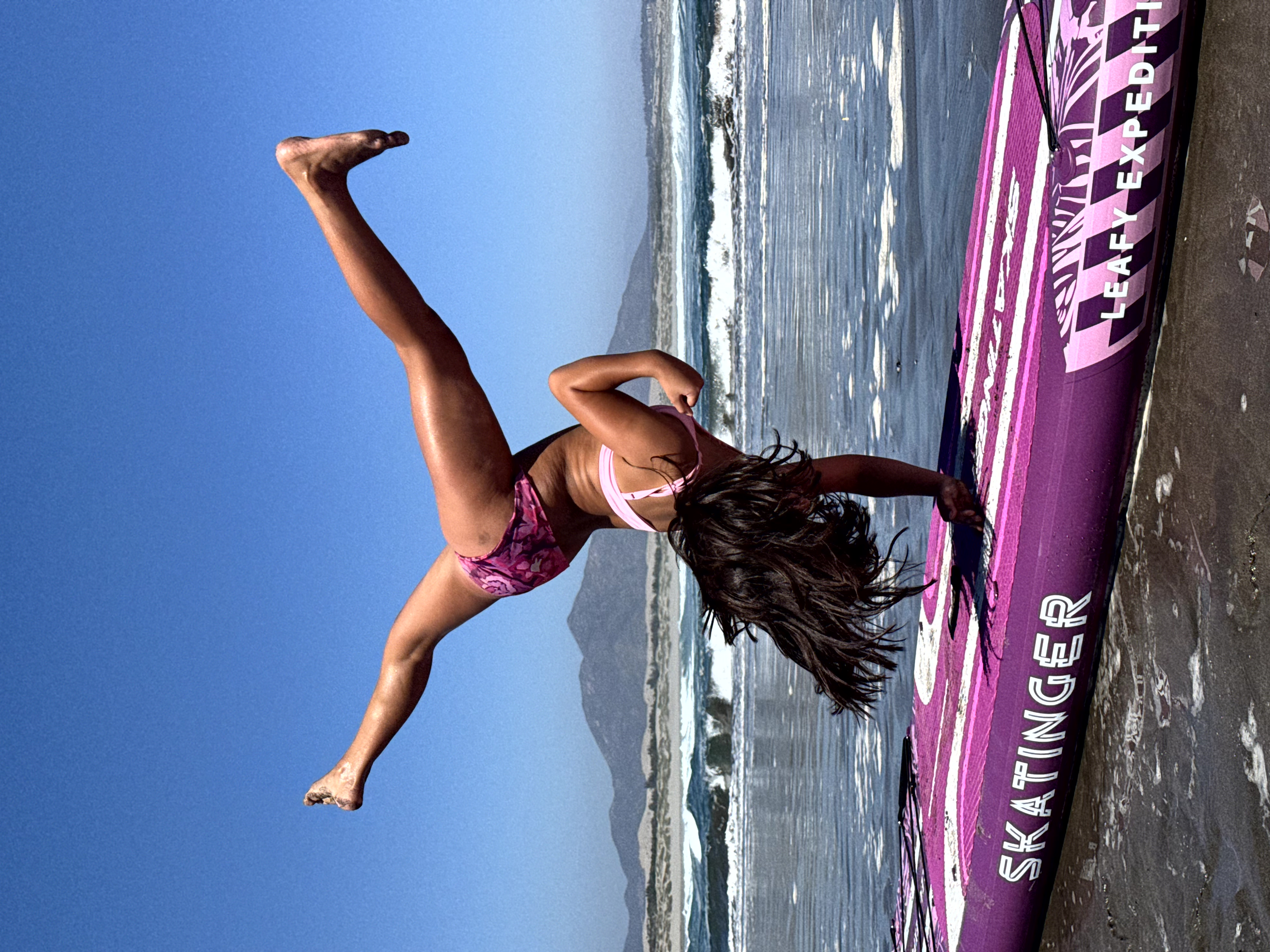 Kobieta w stroju różowym skacze nad fioletową deską SUP na plaży.