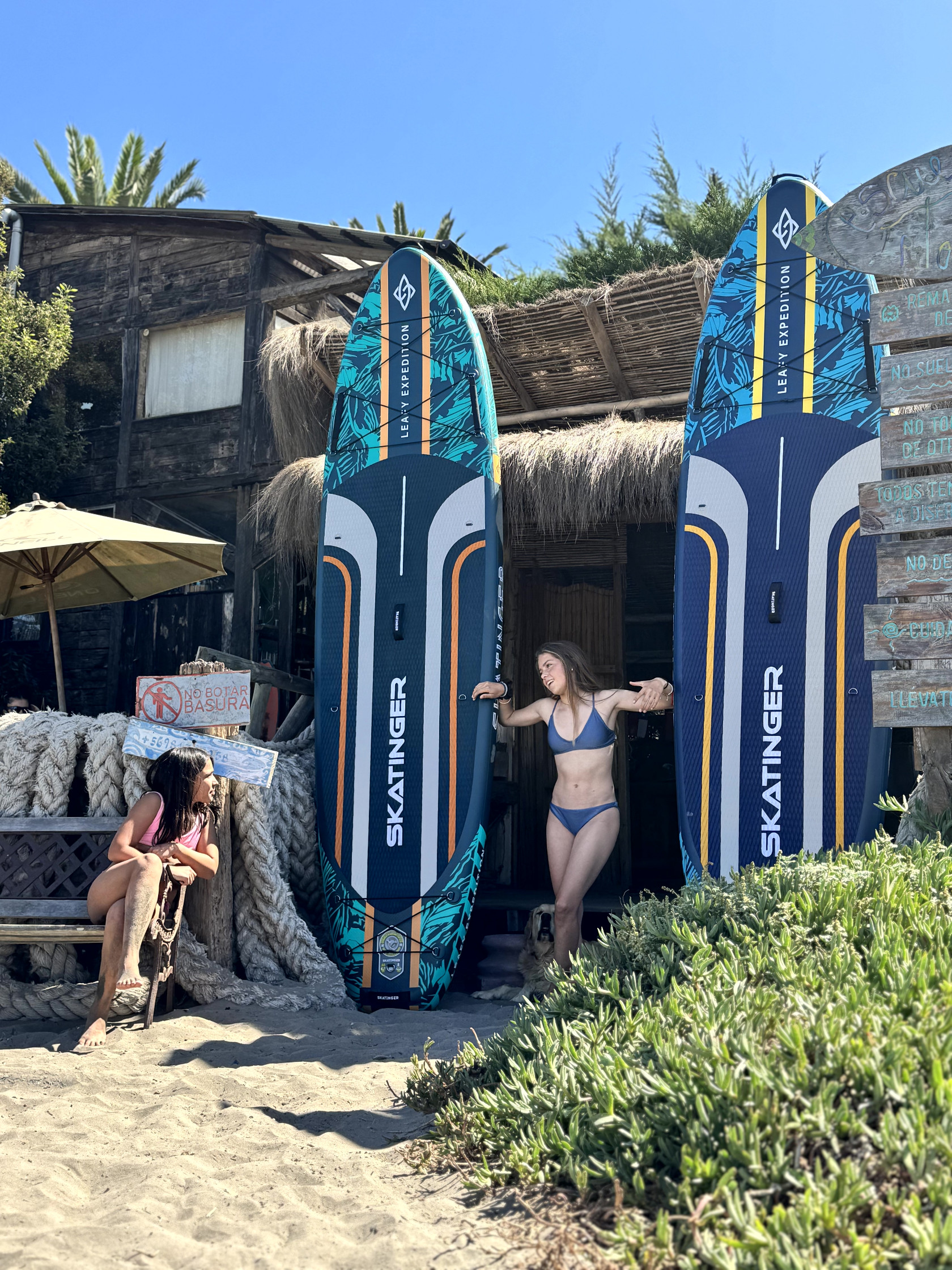 Dwie kobiety i pies stoją przy dwóch deskach surfingowych przed plażową chatką.