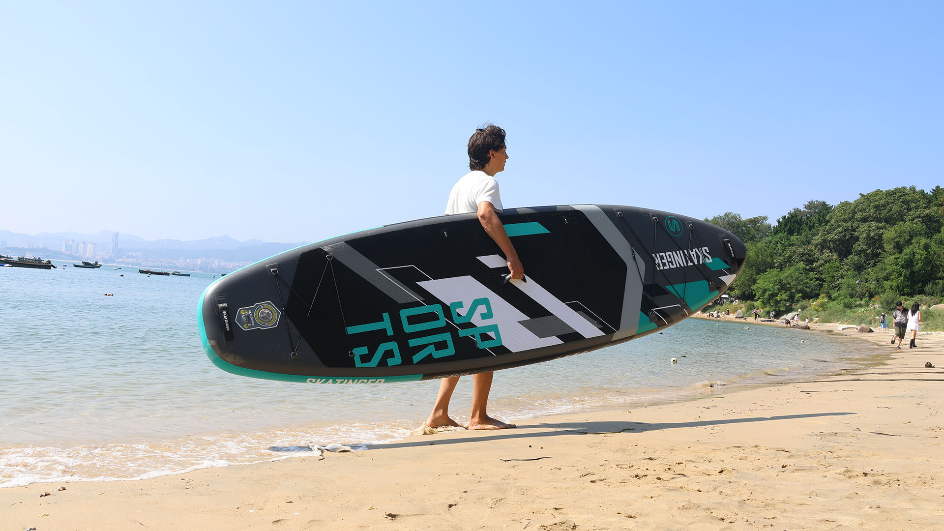 Mężczyzna niesie czarno-morską deskę surfingową na plaży z morzem i drzewami w tle.
