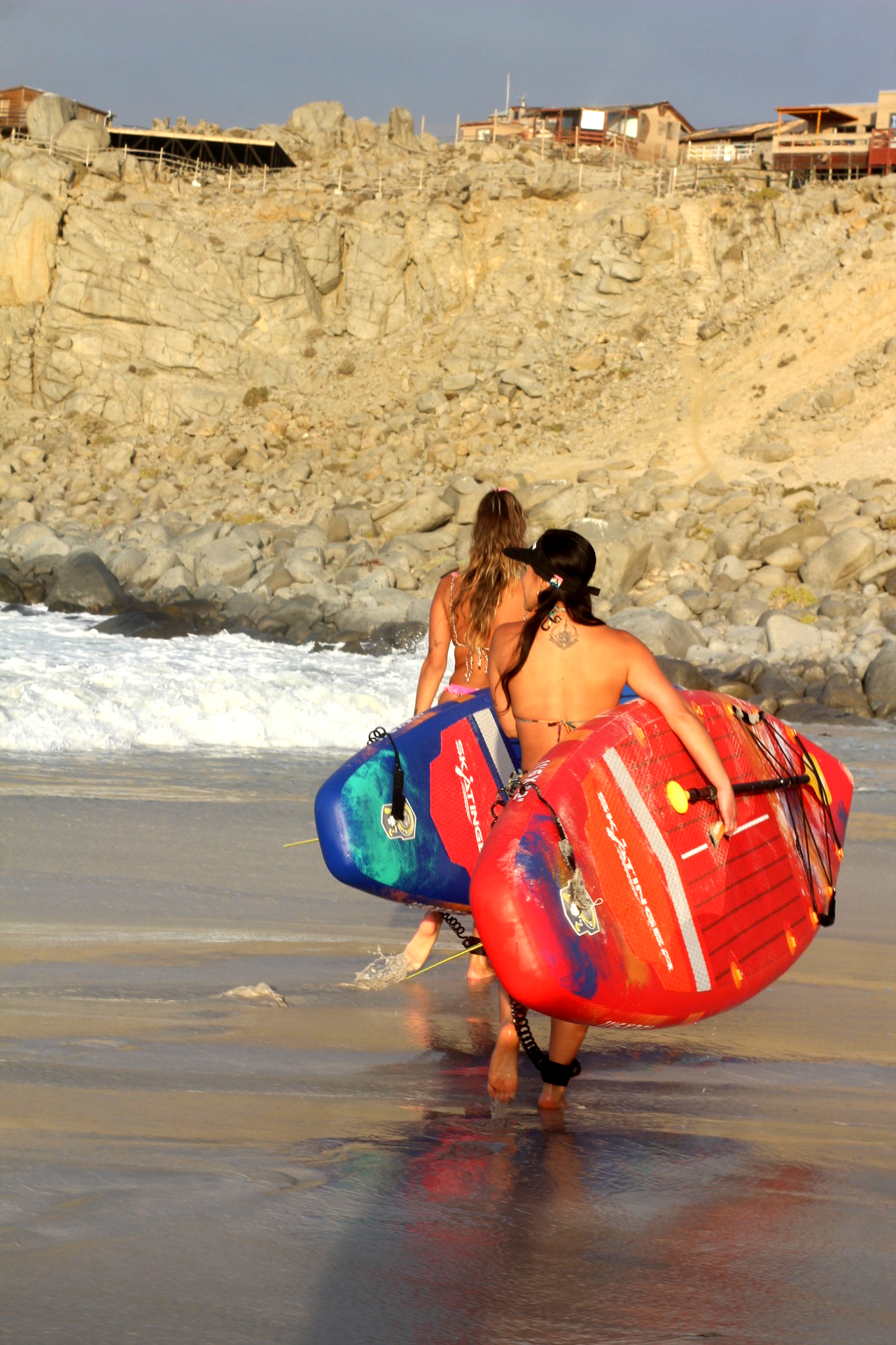 Dwie kobiety idą plażą niosąc deski surfingowe. Skaliste klify z budynkami w tle.