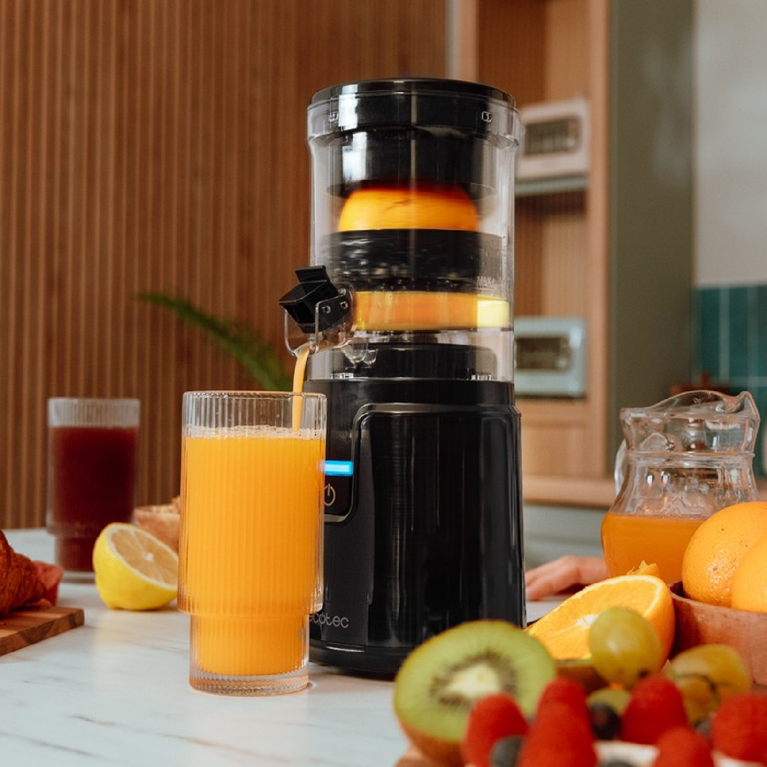 Una licuadora exprime naranjas. Hay vasos de jugo y frutas cerca.