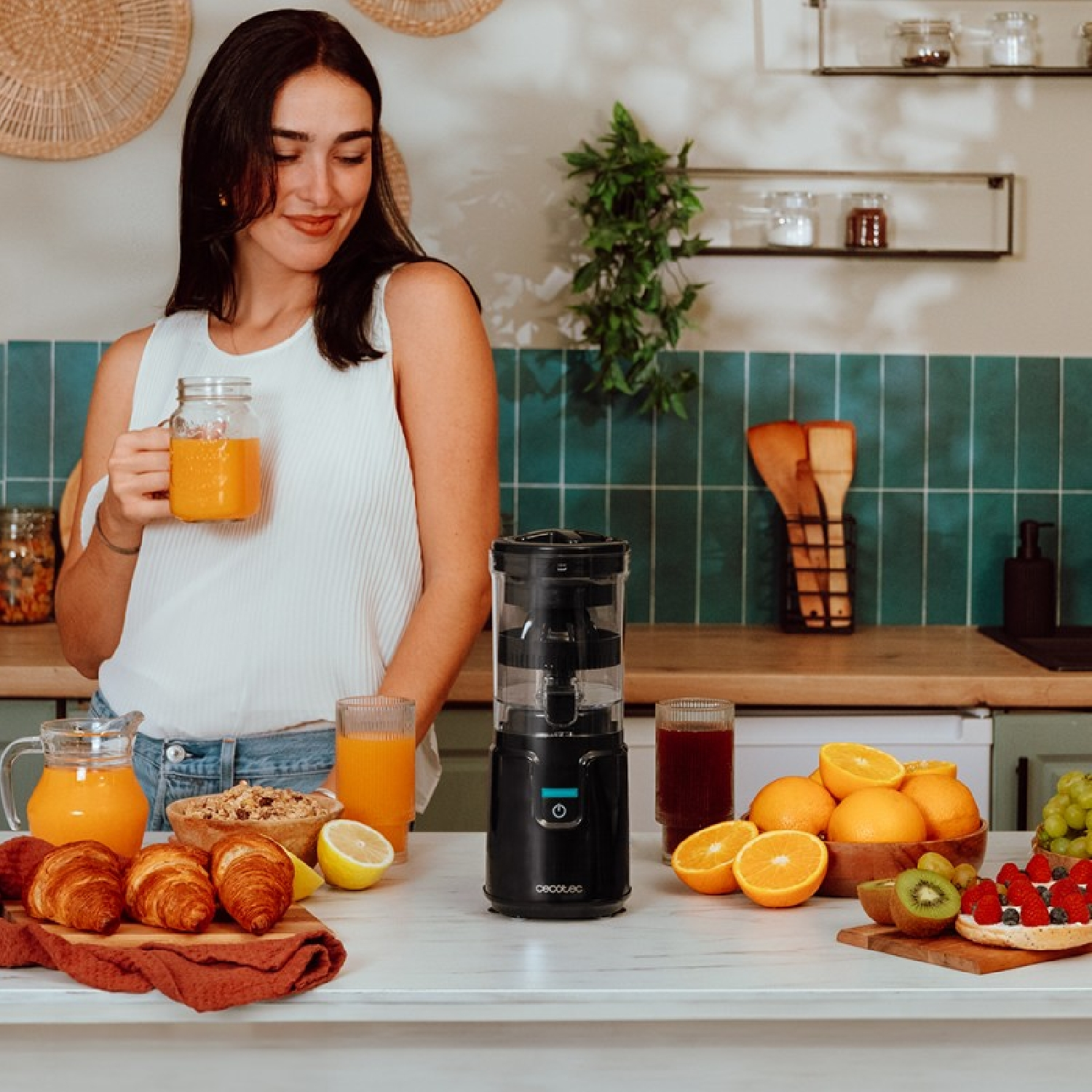 Una mujer sostiene jugo. Una licuadora, frutas y pasteles en el mostrador.