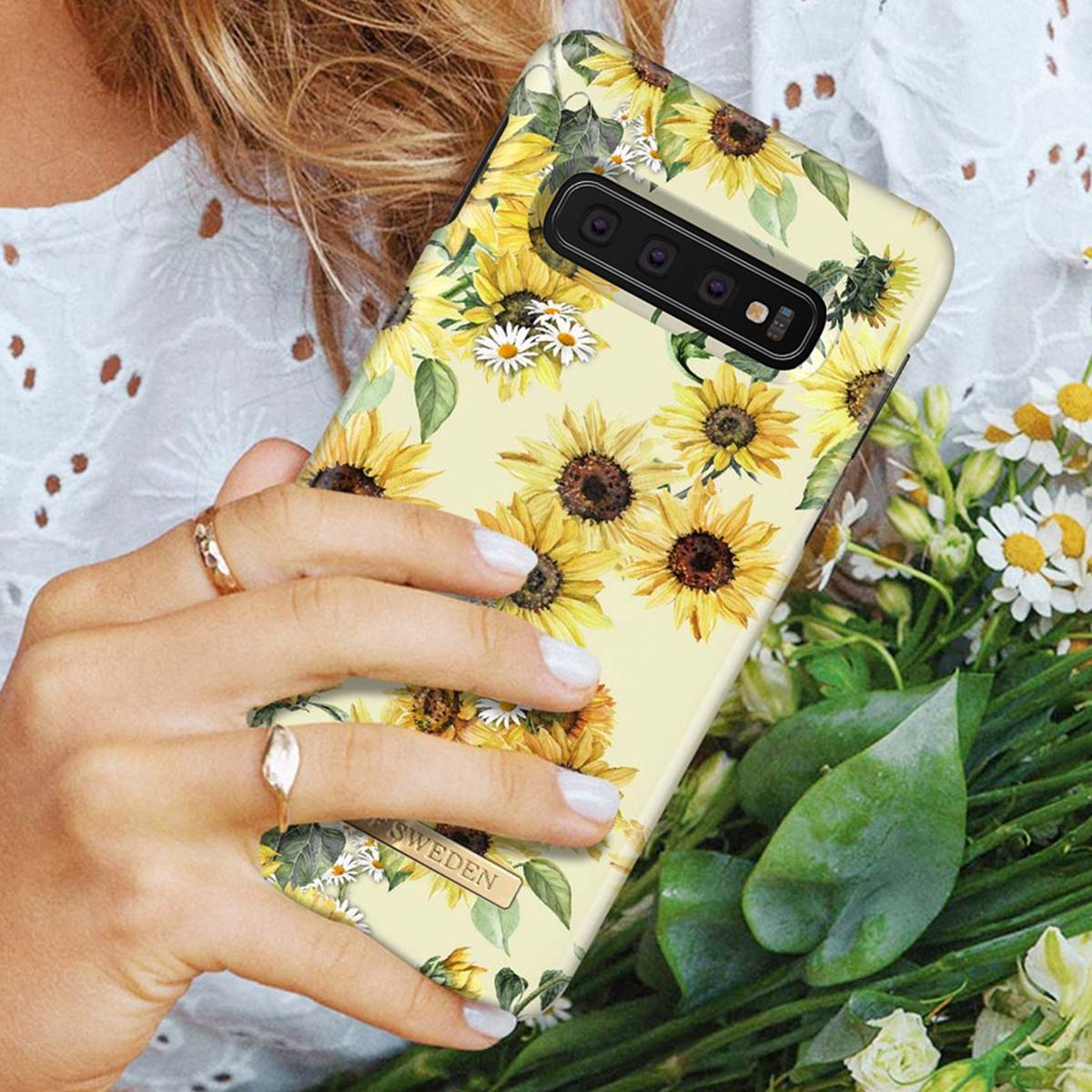Una mano tiene un telefono con motivo di girasoli. Foglie verdi in primo piano.