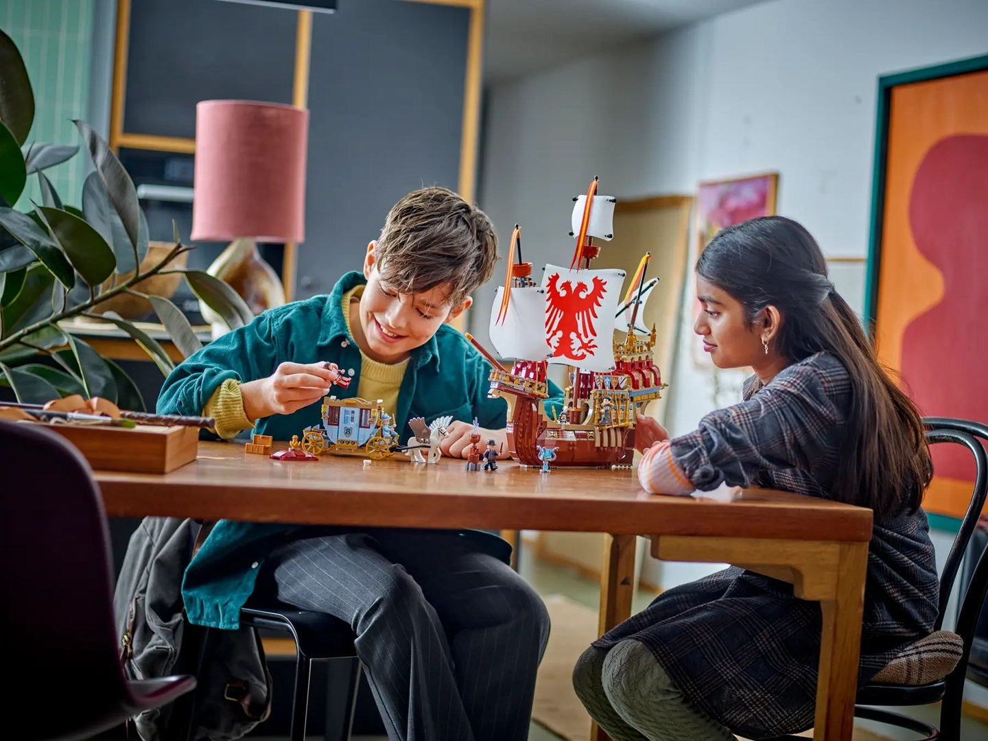 Dwoje dzieci buduje statek i powóz z klocków Lego przy drewnianym stole.