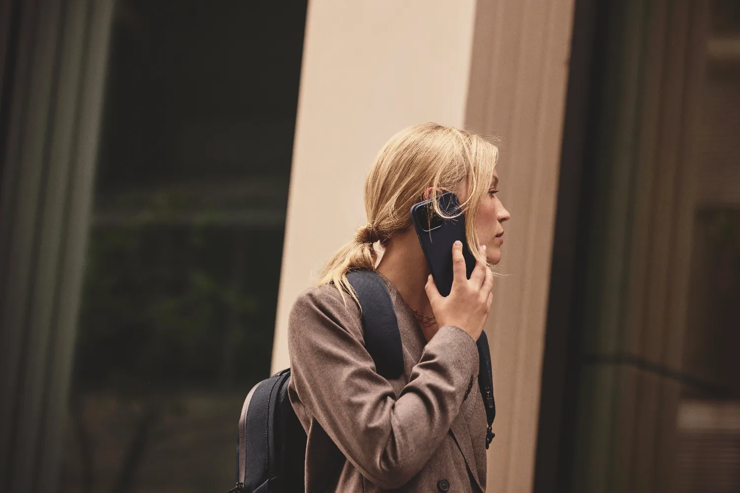 Blondynka rozmawia przez telefon, nosi brązowy płaszcz i plecak.