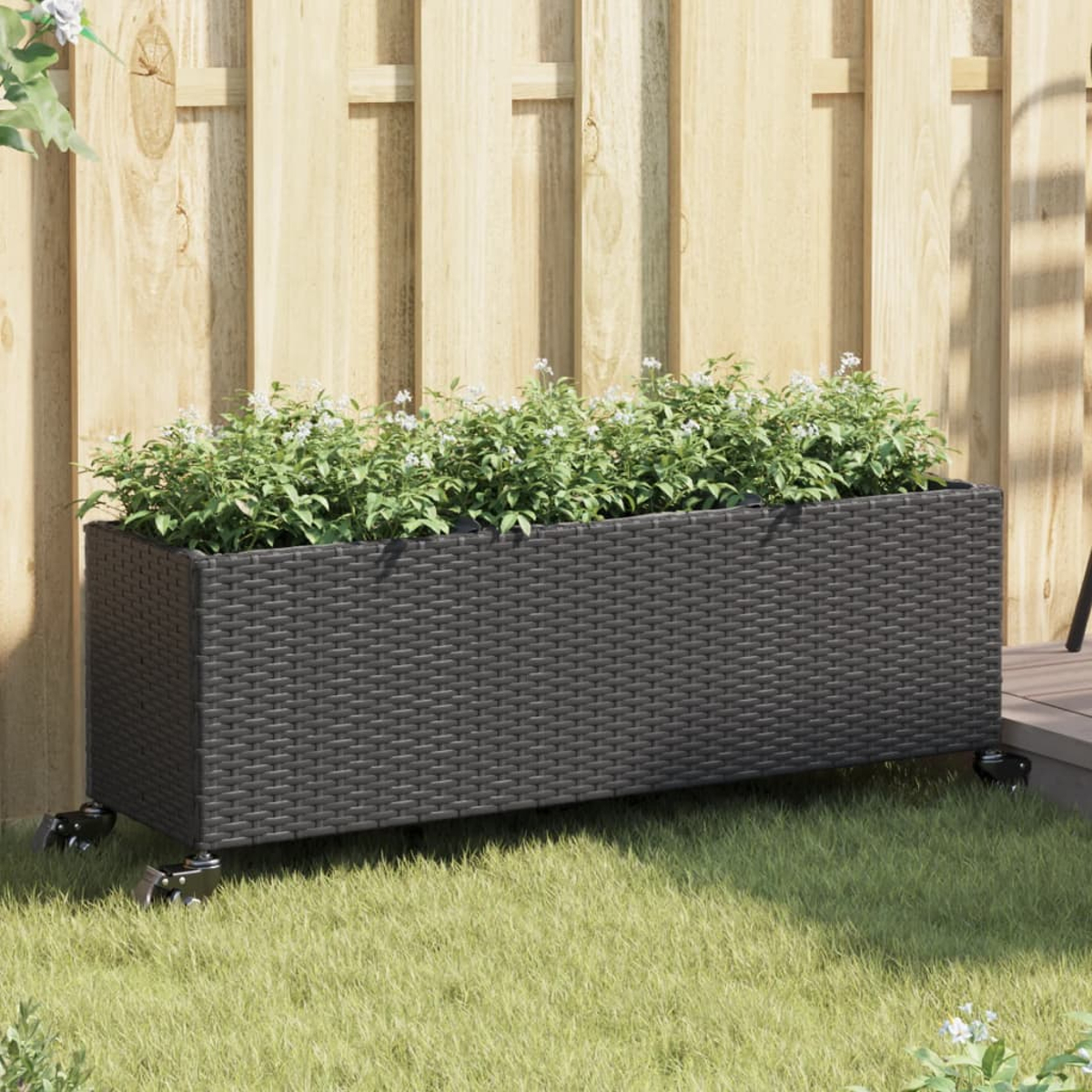 Fioriera in rattan grigio scuro con ruote e piante verdi, davanti a una recinzione di legno.