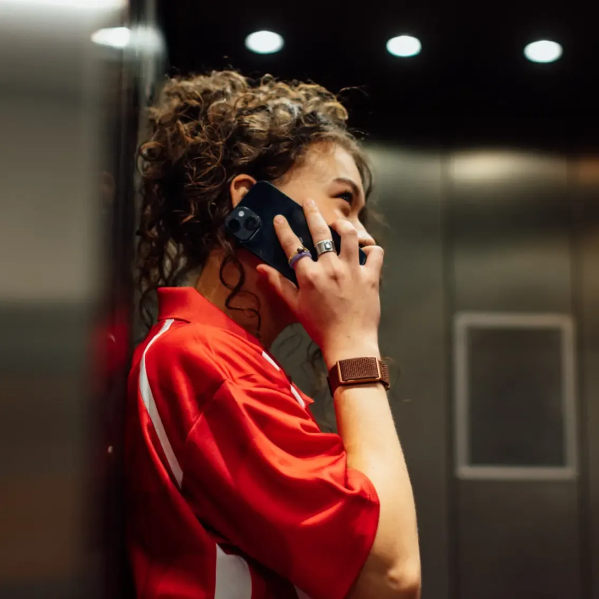 Mujer joven con pelo rizado habla por teléfono en ascensor. Viste polo rojo.