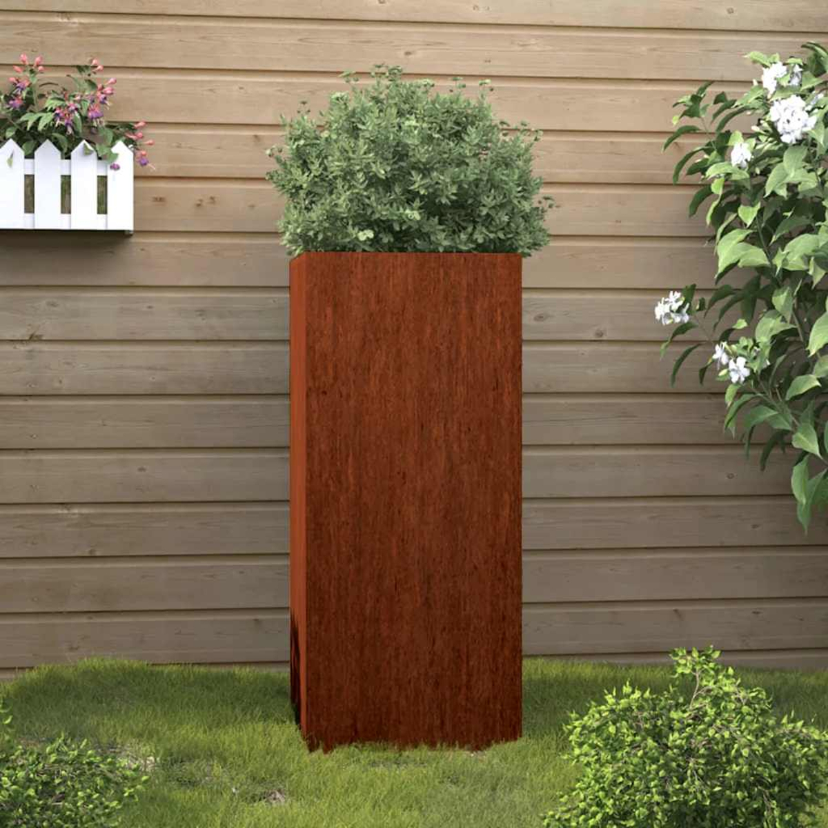 Ein hoher, rostfarbener Pflanzkübel mit grünem Busch auf Gras vor Holzwand.
