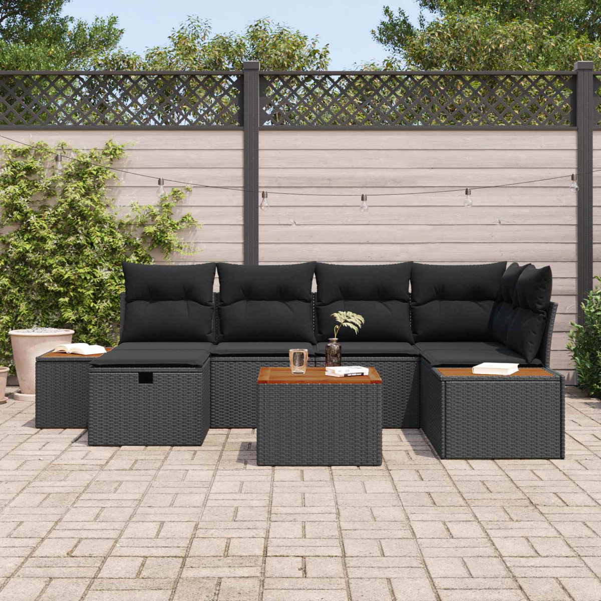 Graues Rattan-Sectioal-Sofa mit schwarzen Kissen auf einer gepflasterten Terrasse, mit Zaun und Pflanzen.