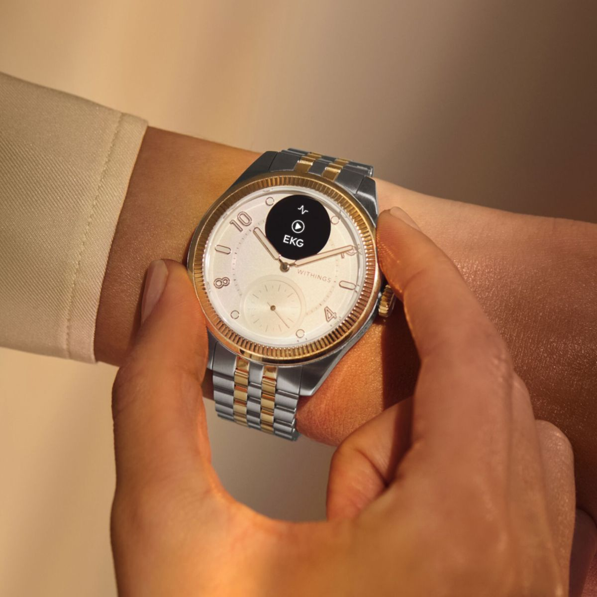 Osoba nosi zegarek z srebrną i złotą bransoletą. Na ekranie widnieje EKG.