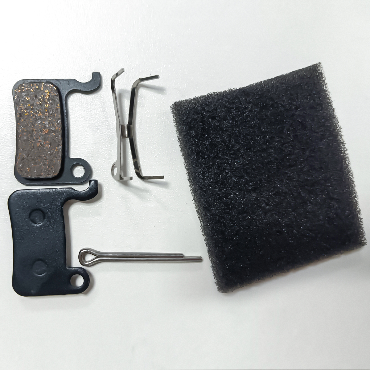 Dwa klocki hamulcowe rowerowe, sprężysty klips, sworzeń i czarny blok pianki.
