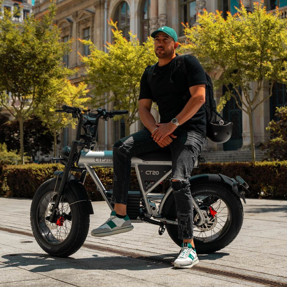 Un uomo con la barba seduto su una e-bike argentata davanti a un edificio con alberi.