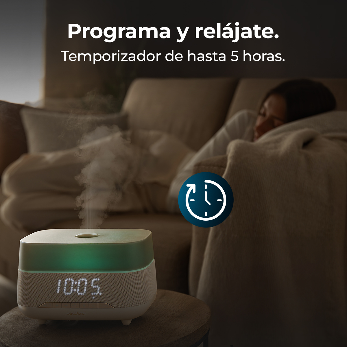 Un diffusore con luce verde e icona del timer su un tavolo. Una persona dorme sul divano.