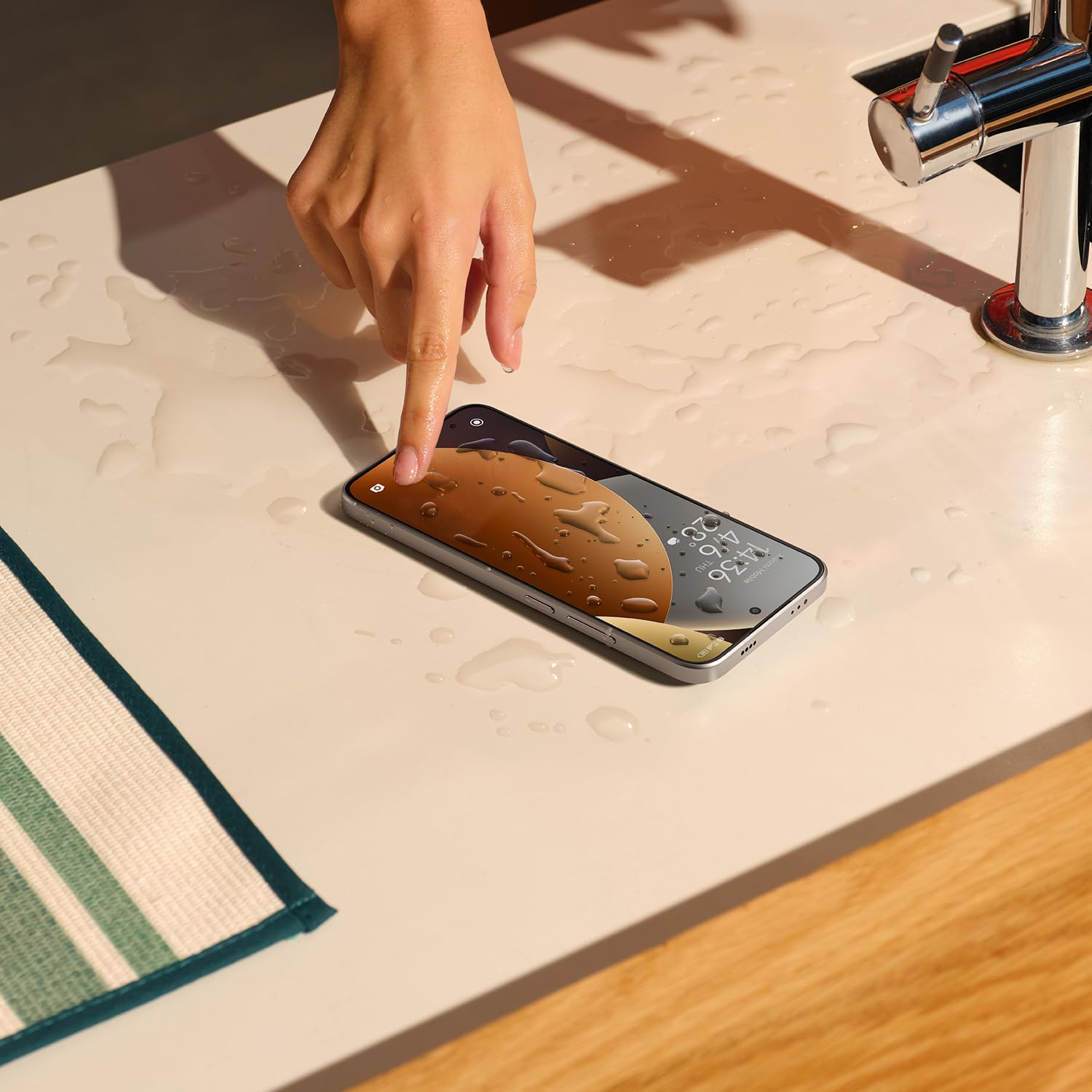 Eine Hand berührt ein Smartphone auf einem nassen Tisch neben einem Wasserhahn.
