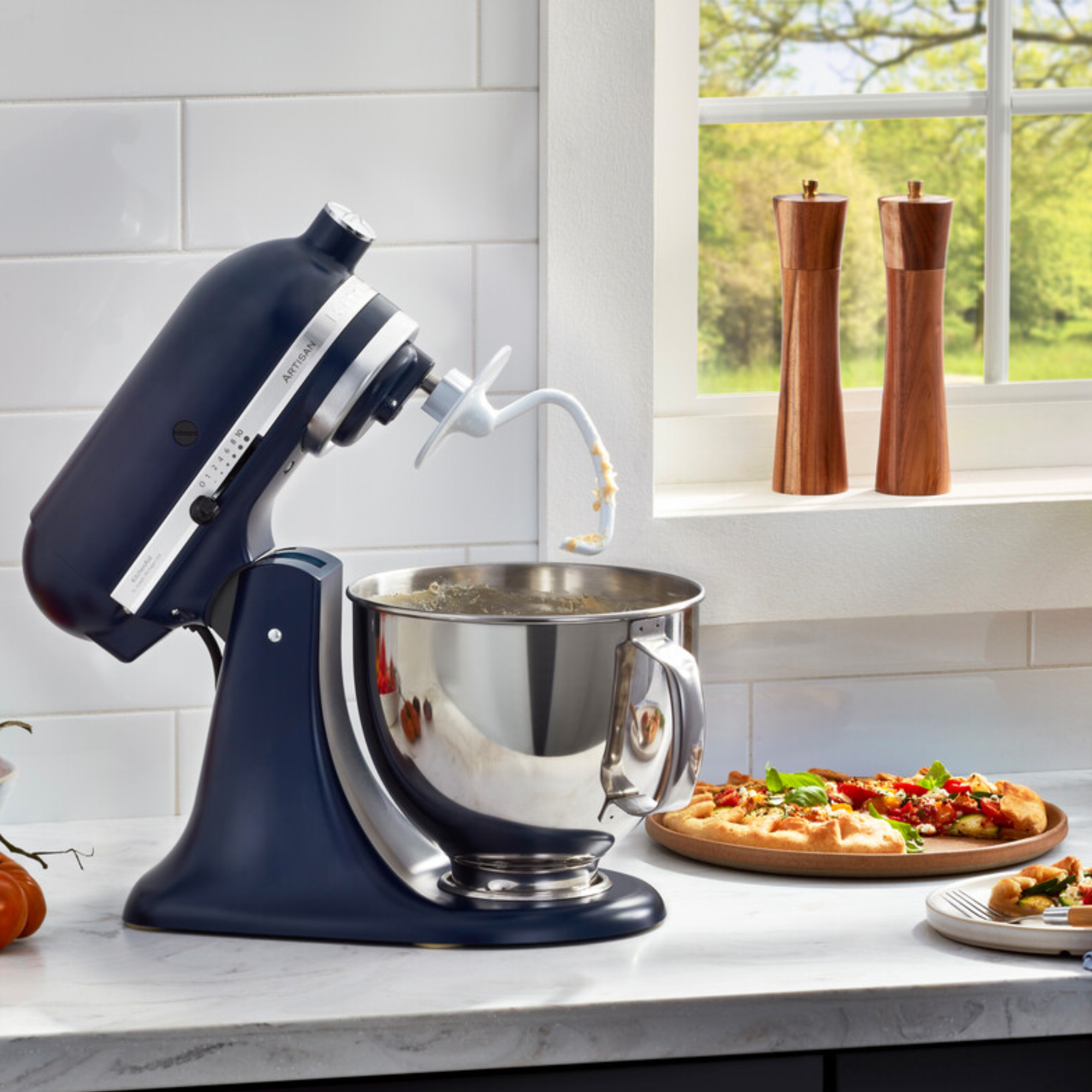 Dunkelblauer Küchenmixer mit Teighaken in Edelstahlschüssel, daneben Pizza.