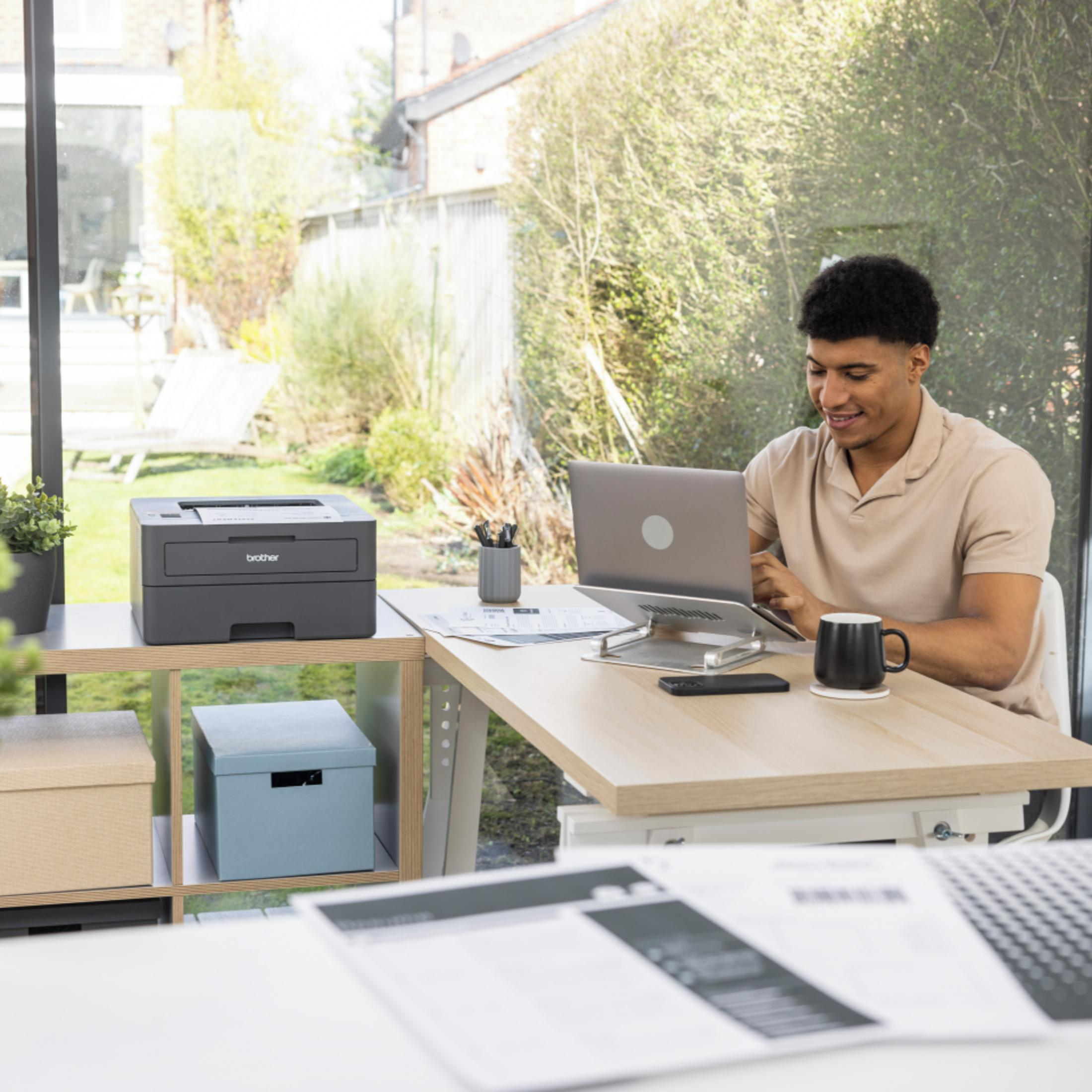 Mężczyzna pracuje przy laptopie, drukarce i materiałach biurowych na biurku z widokiem na ogród.