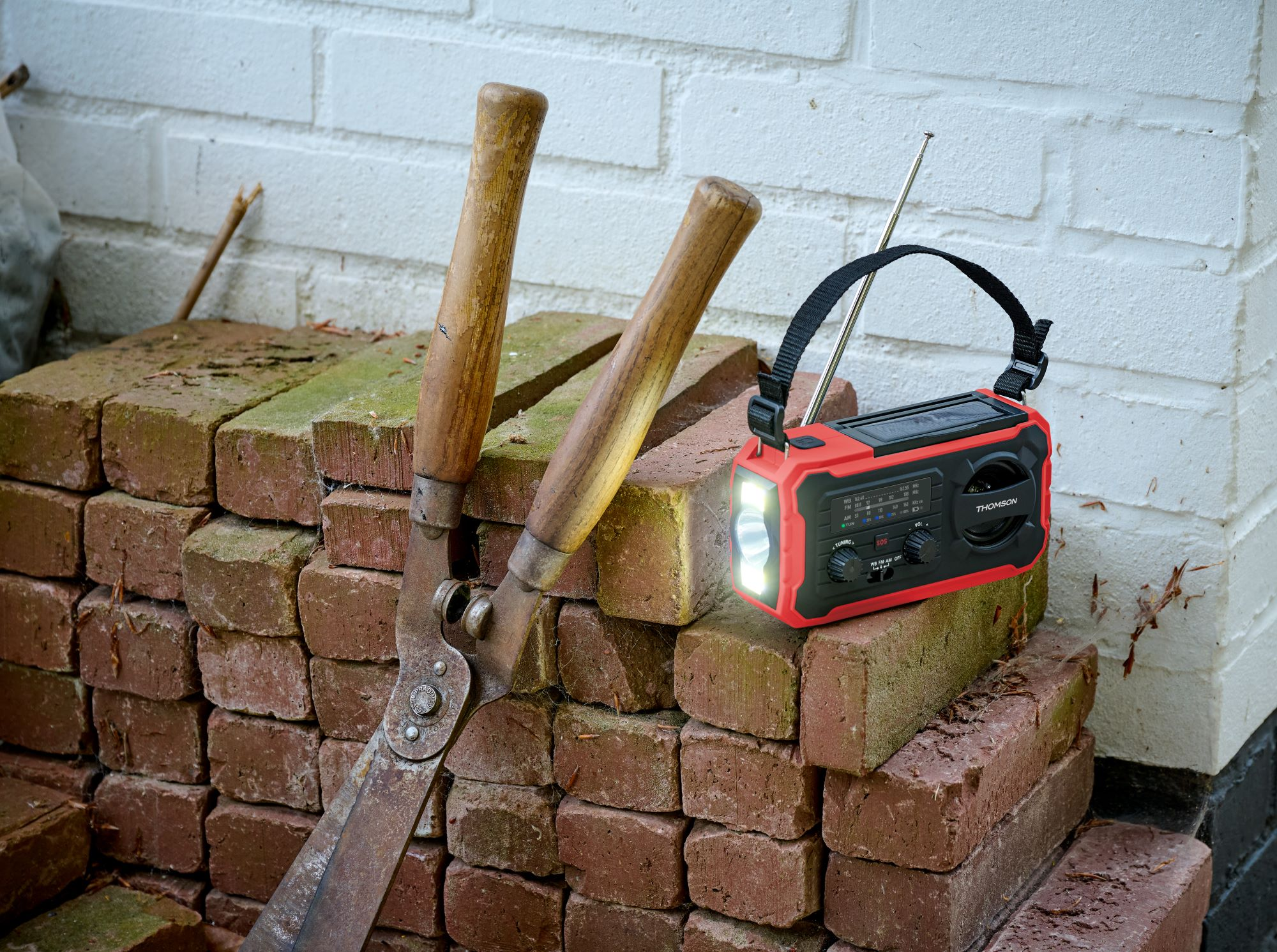 Una radio rossa con luce è su mattoni accanto a cesoie da giardino.