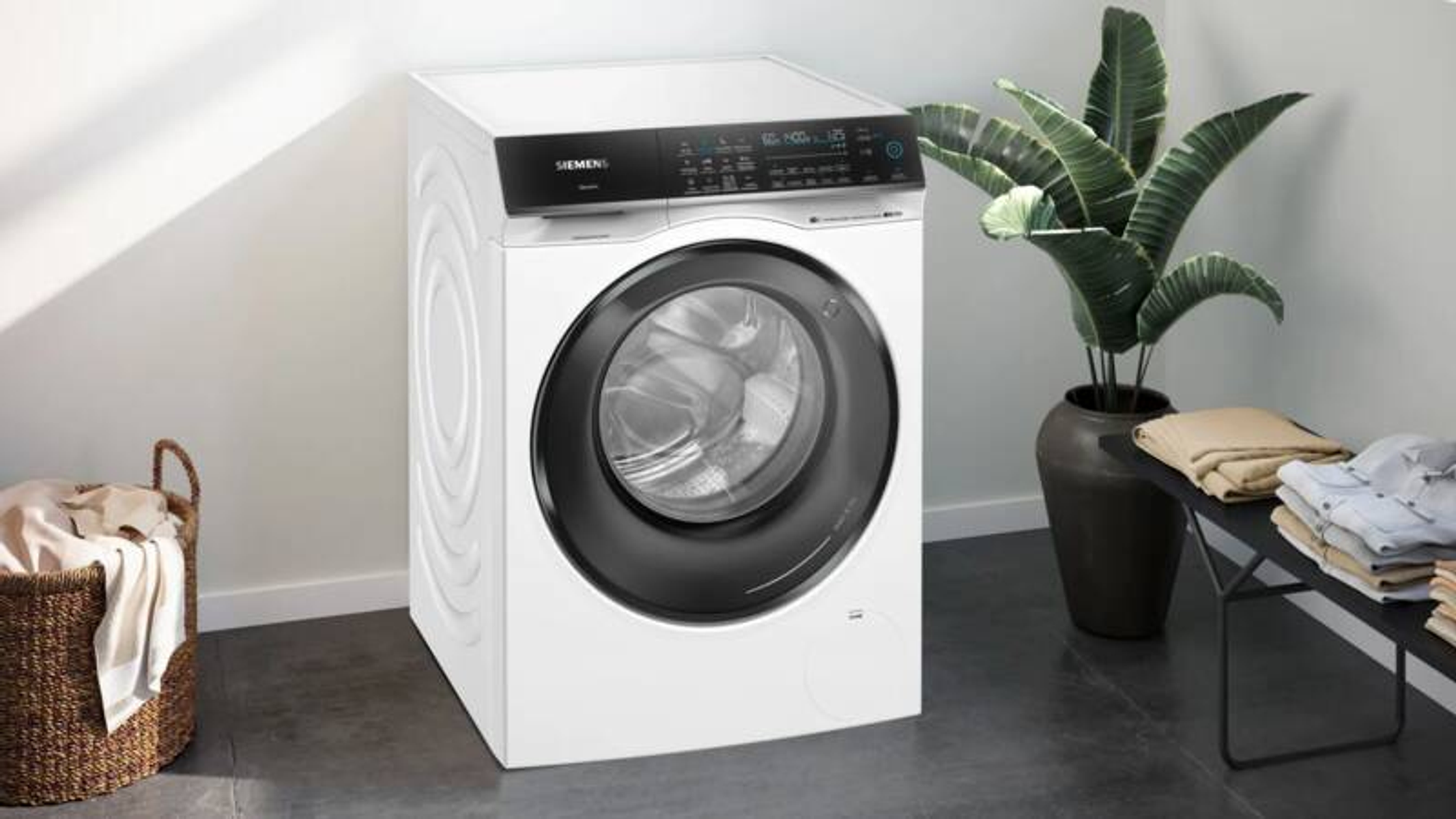 Eine weiße Siemens Waschmaschine steht neben einem Wäschekorb und einer Pflanze.