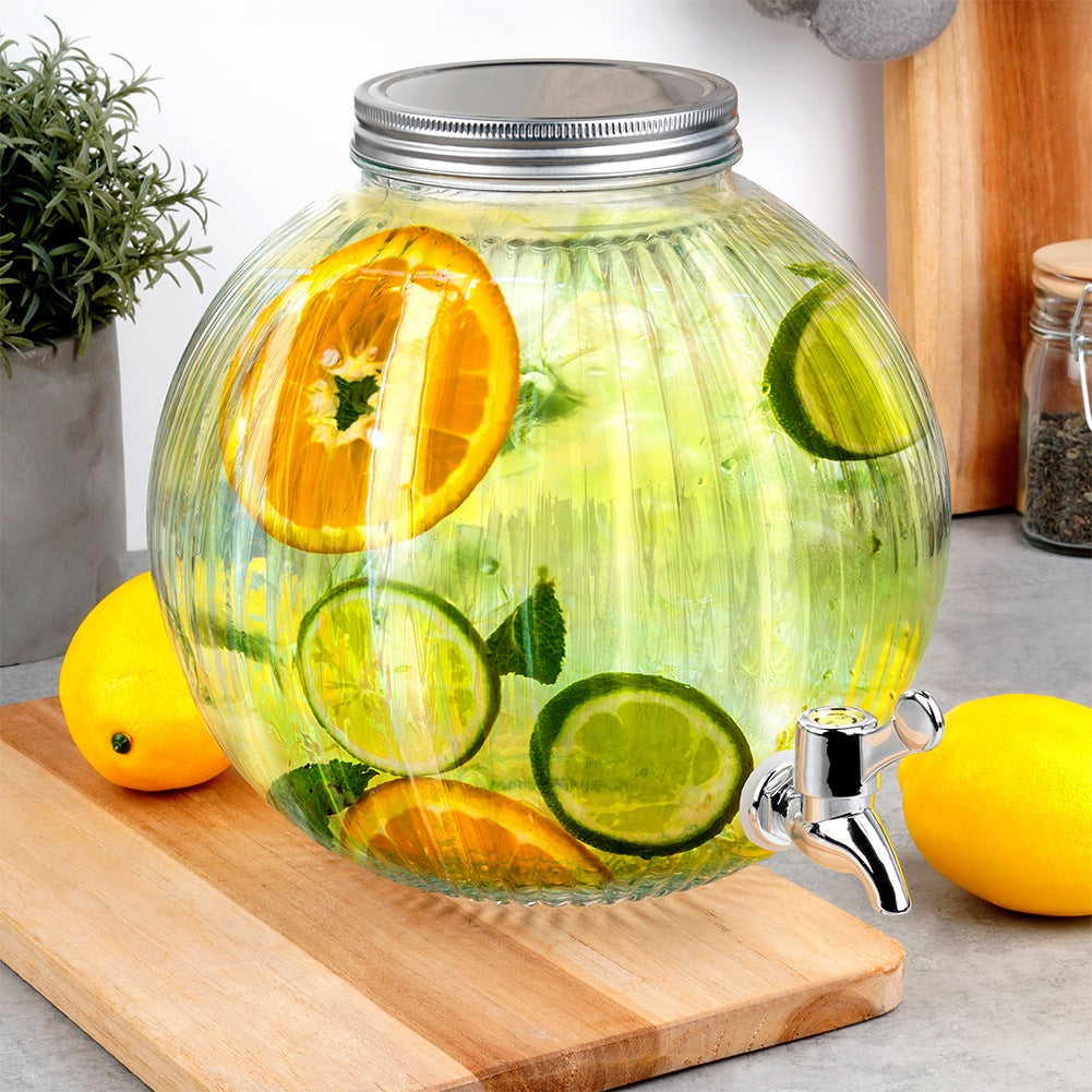 Dispensador de vidrio con fruta y grifo sobre tabla de madera.