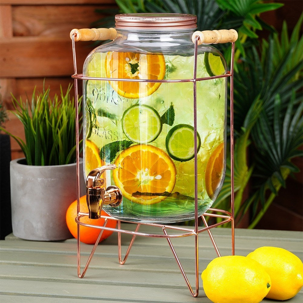Dispensador de bebidas con rodajas de naranja y lima, sobre un soporte de cobre.