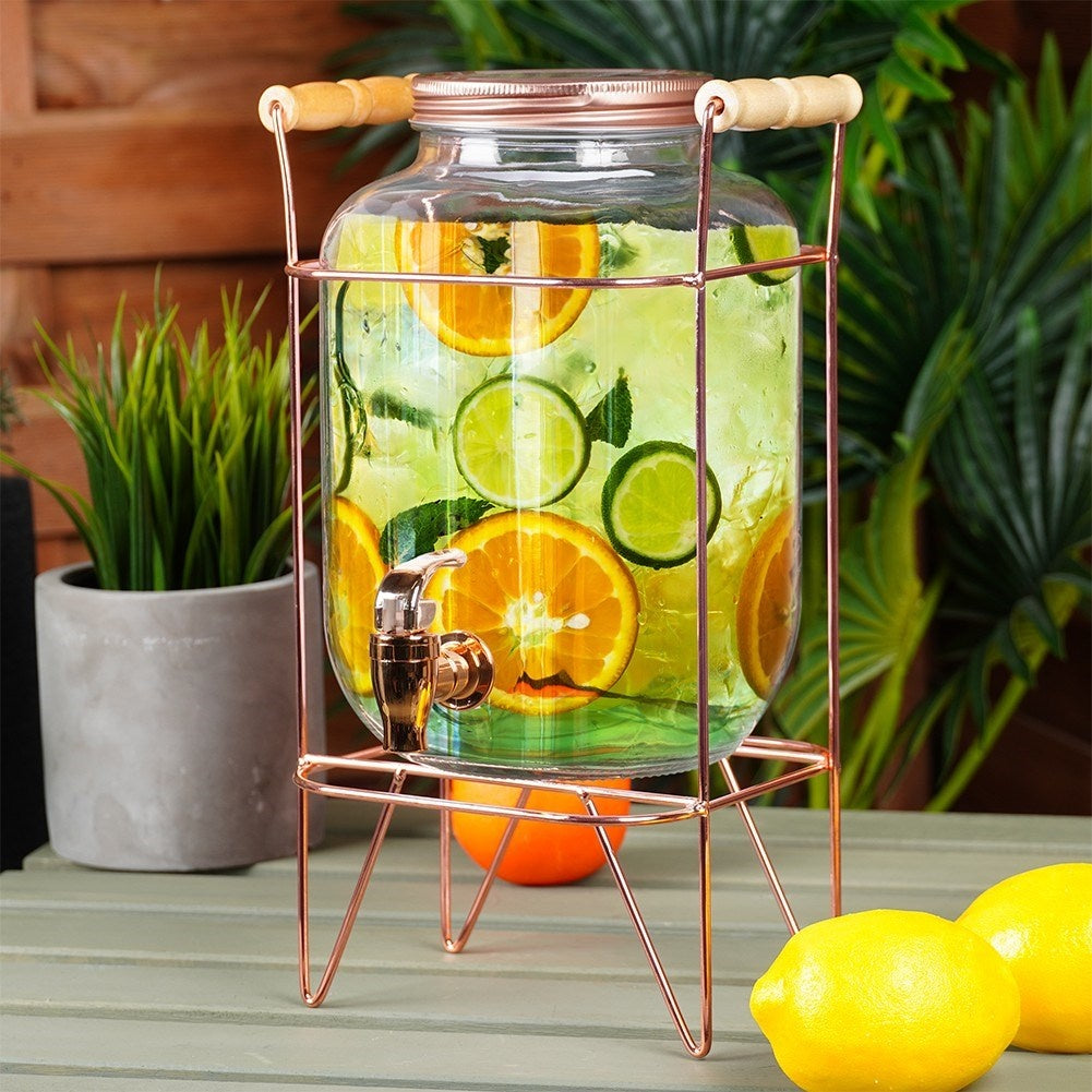Un dispensador de vidrio con grifo, lleno de agua, hielo y rodajas cítricas, sobre un soporte de cobre.