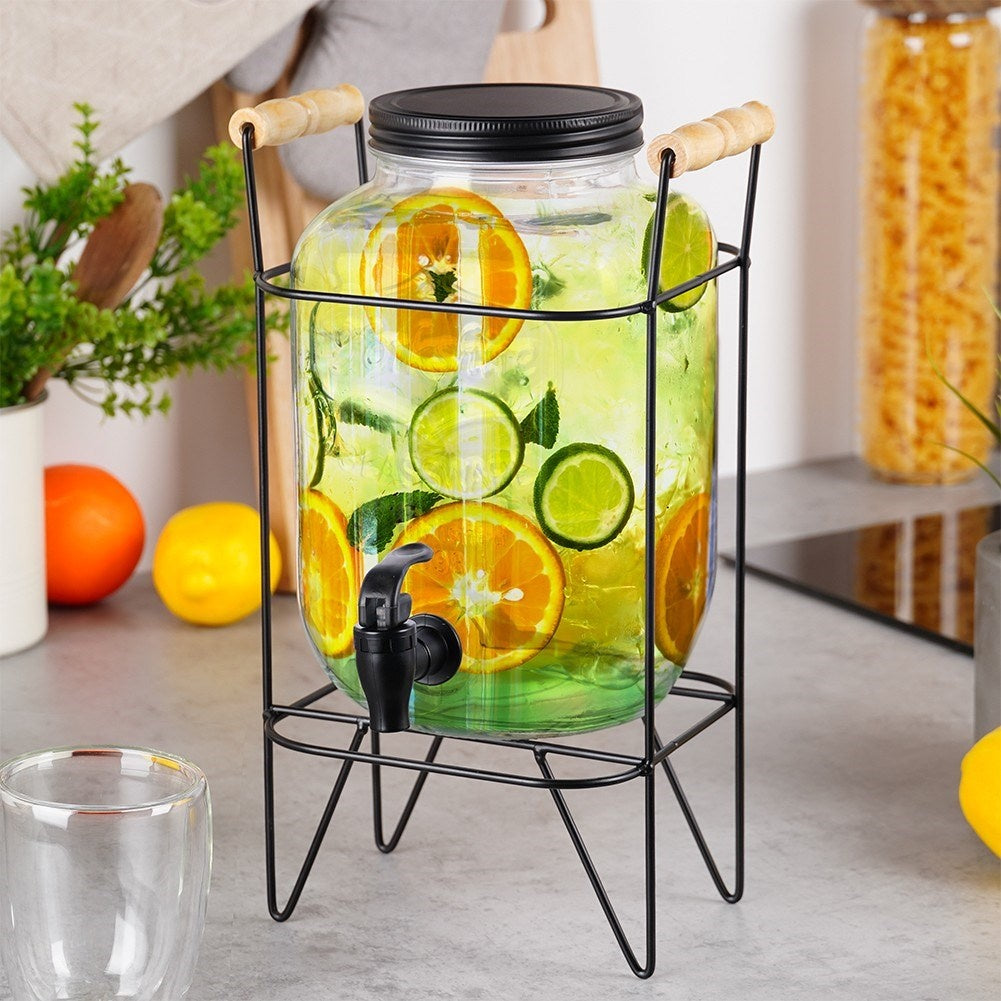 Dispensador de bebidas de vidrio con naranjas, limas y menta en un soporte.