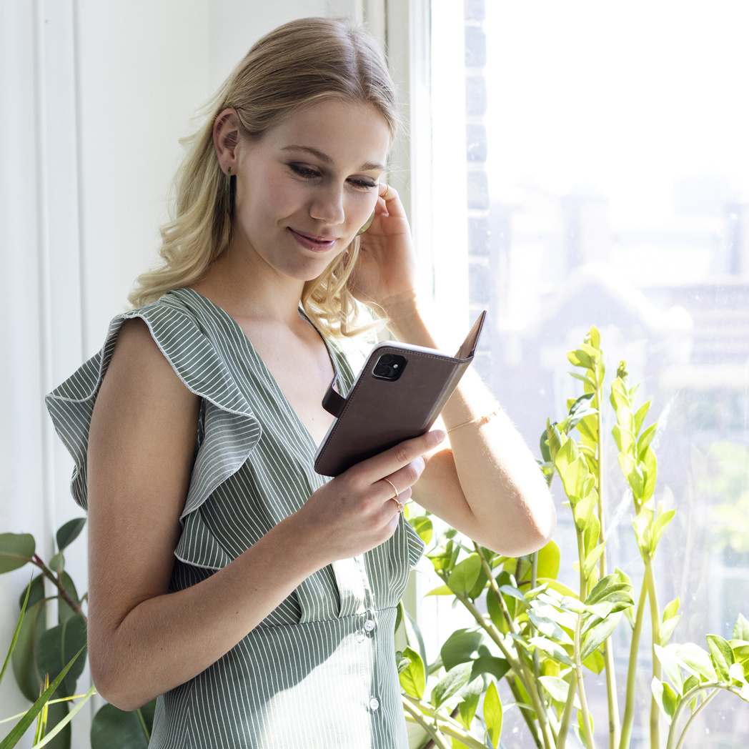 Une femme blonde tient un téléphone près de plantes près d'une fenêtre.